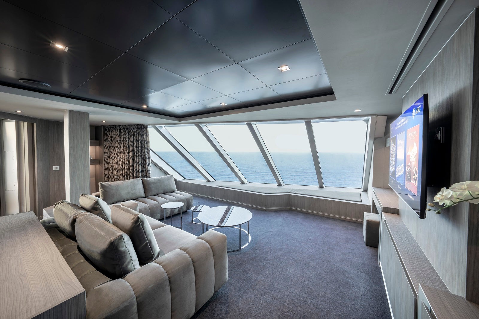 A cruise ship cabin decorated in shades of gray has a wall of windows and a view of the ocean