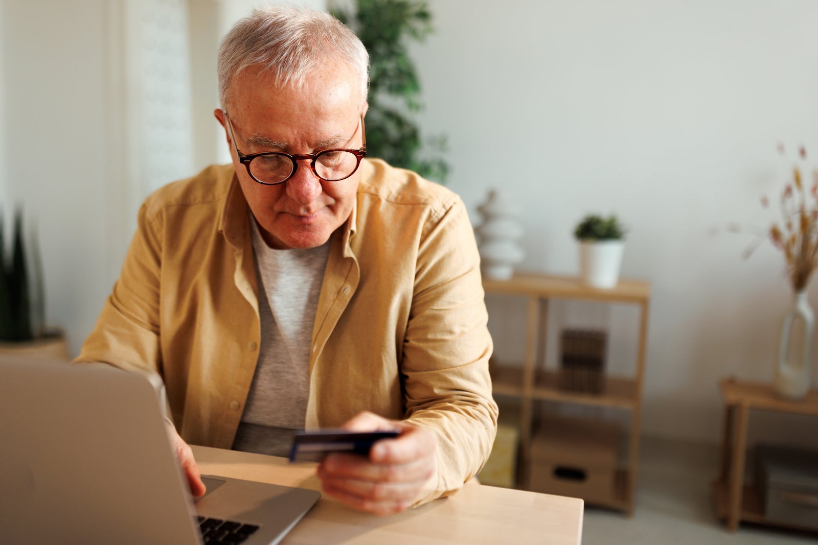 a senior man sits at a kitchen table, holding a credit card while online shopping