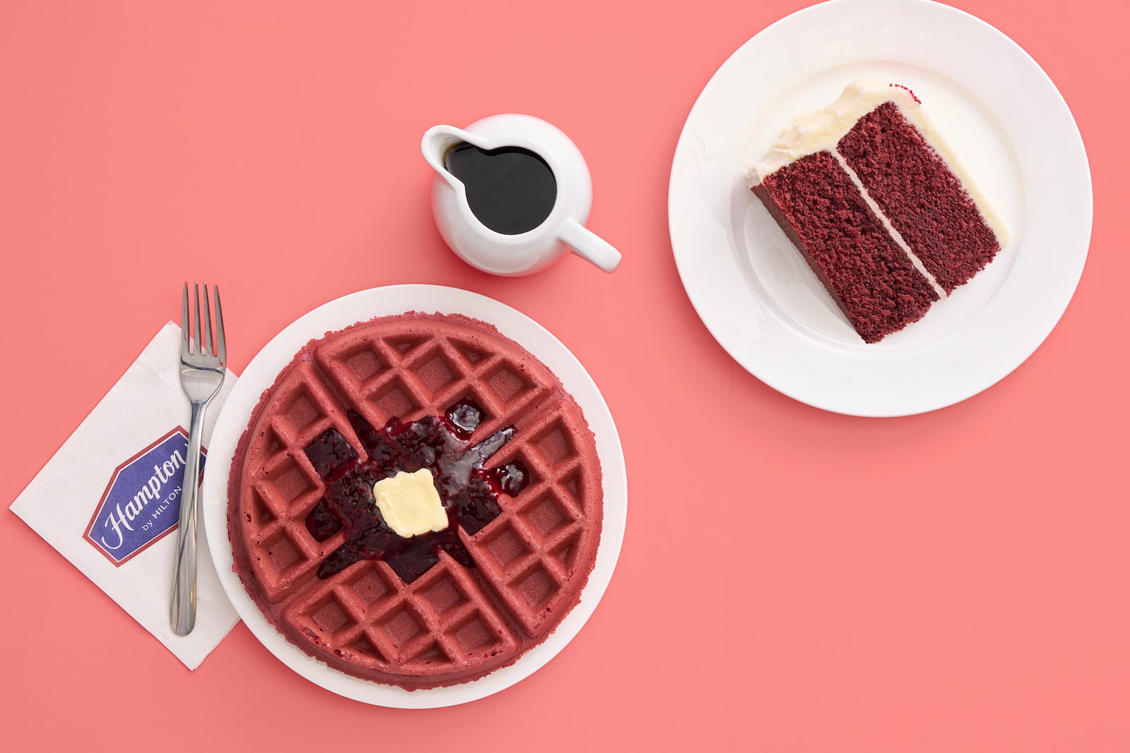 red velvet cake on white plate, red velvet waffle with jam on white plate with fork and jar of syrup on pink table