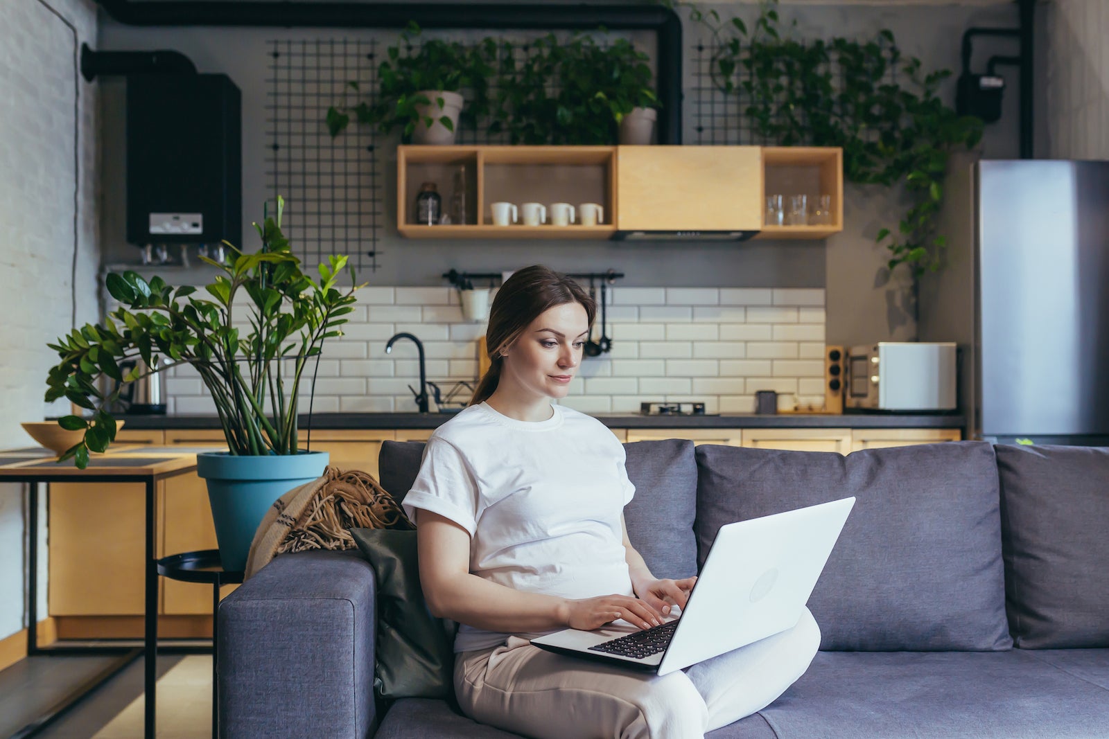 a woman sits on her sofa while using her laptop; a kitchen is visible in the background