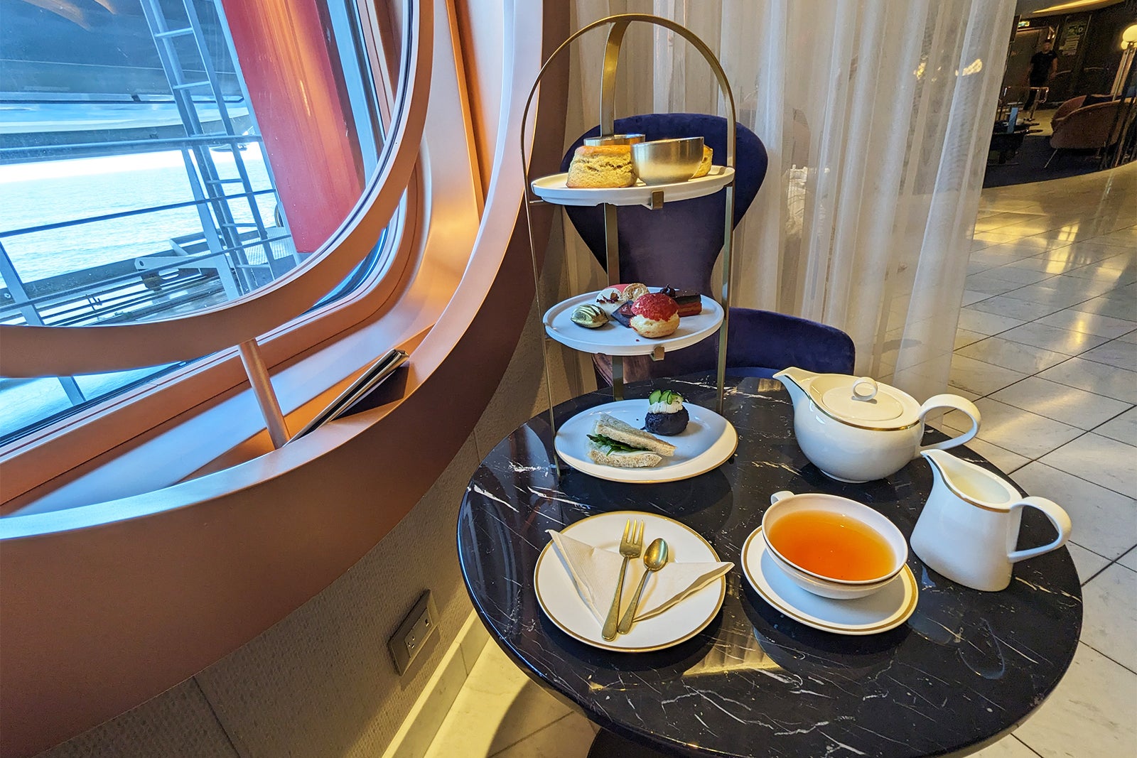 Three-tiered tray of snacks with teapot and cup of tea next to porthole window on cruise ship