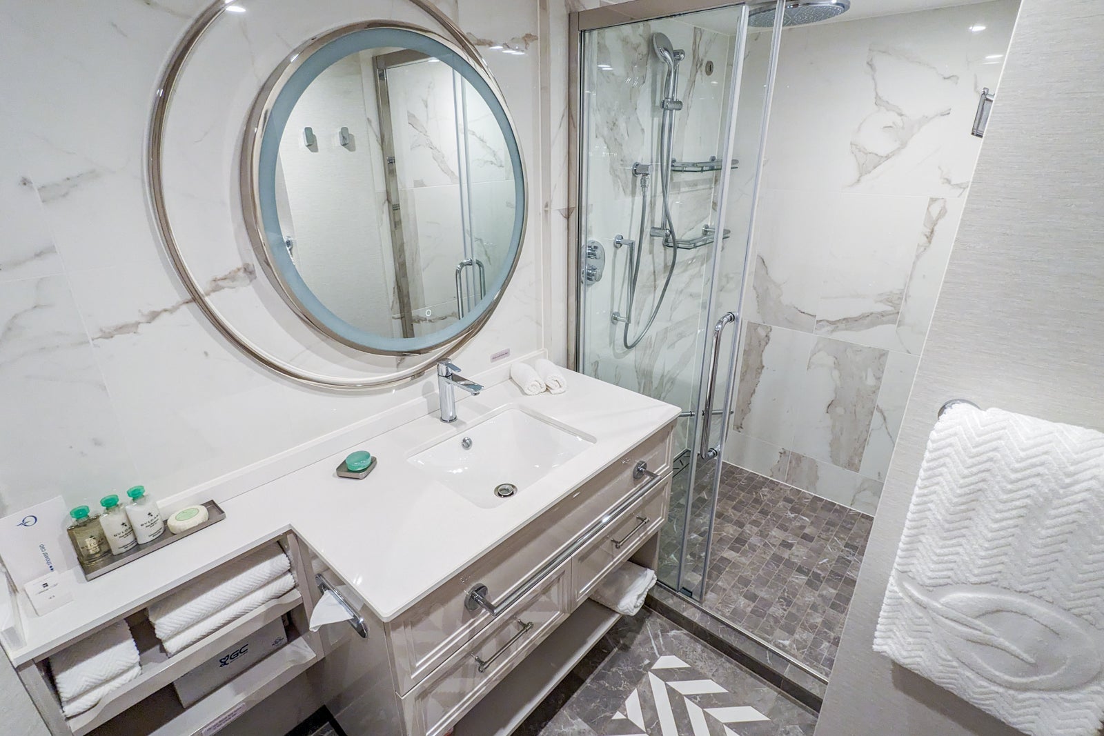 White marble cruise ship bathroom with circular mirror, square sink and glass shower