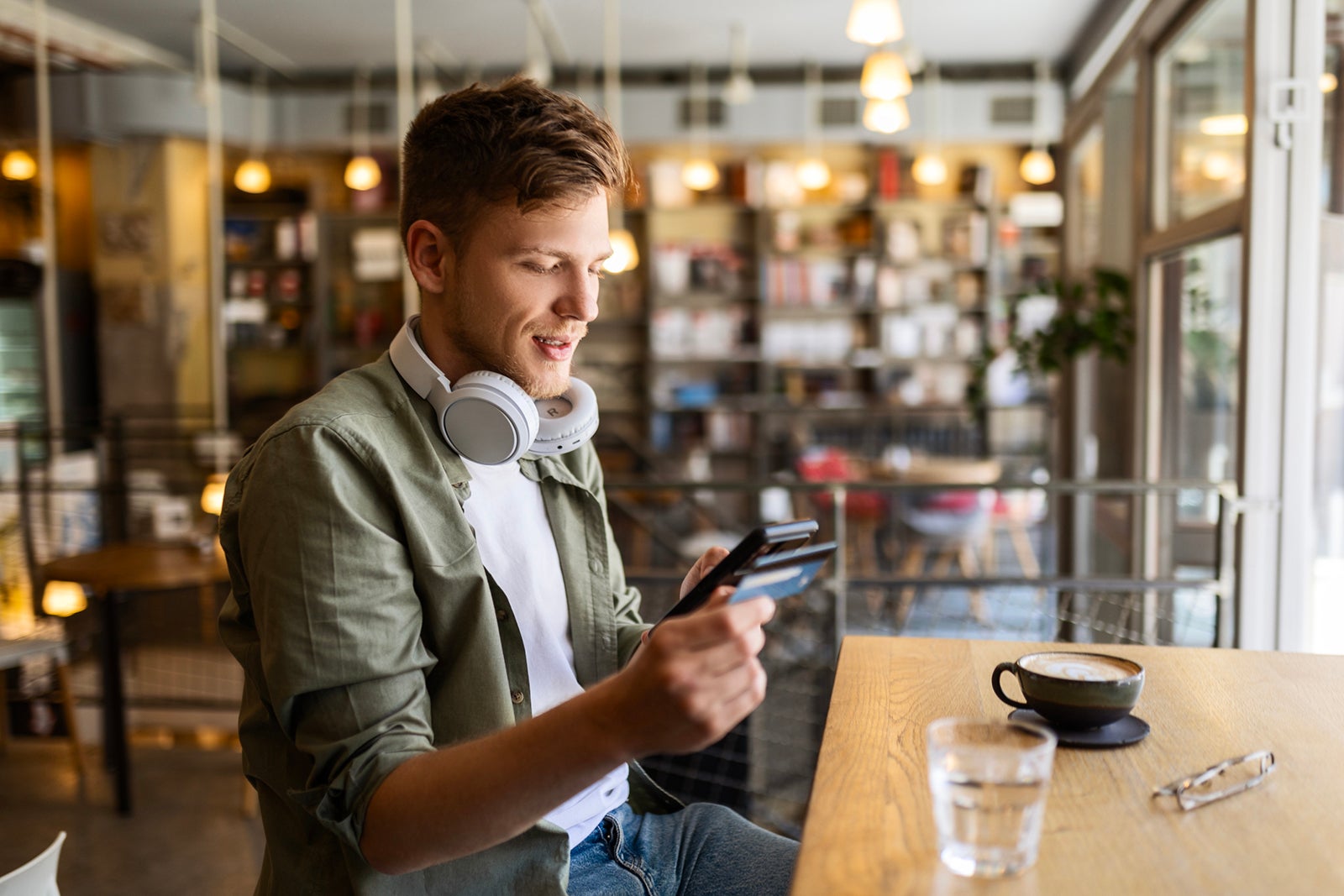 Happy man sitting and drinking coffee at cafe using his phone with credit card