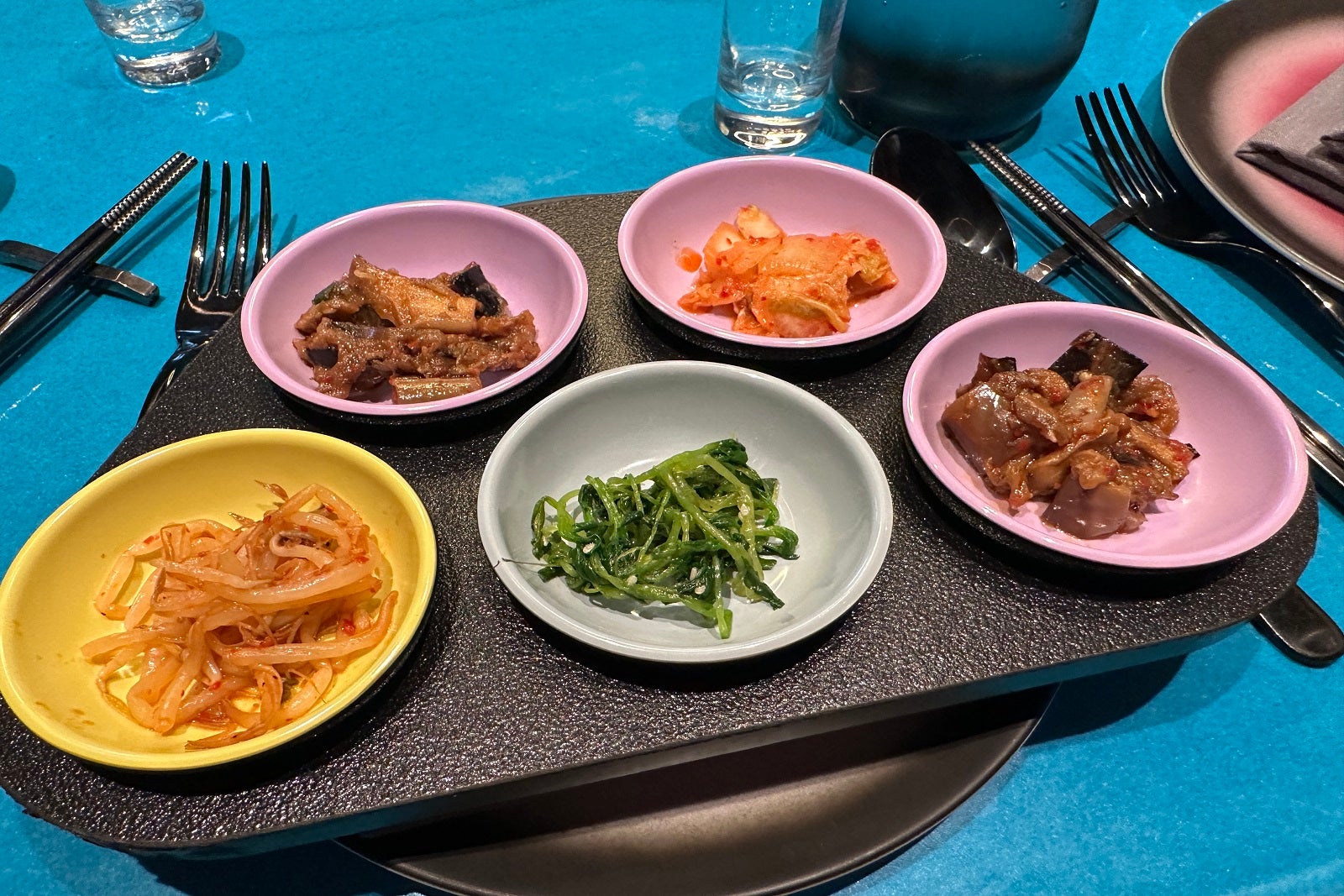 Five small, colorful bowls filled with appetizers sitting on a larger black, rectangular plate on a blue table