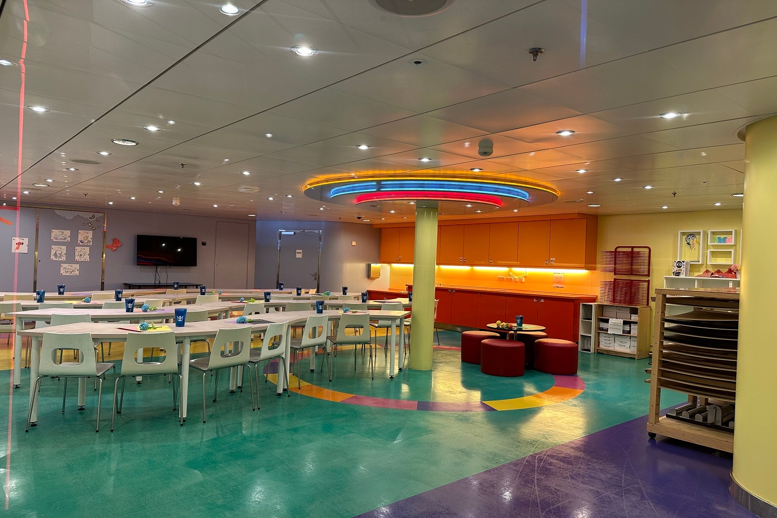 A large room with tables, chairs and colorful overhead lighting in the kids club on a cruise ship