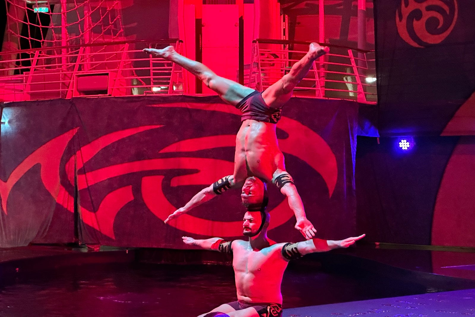 An acrobat sitting on the ground with his arms out and another acrobat balancing on the first acrobat's head