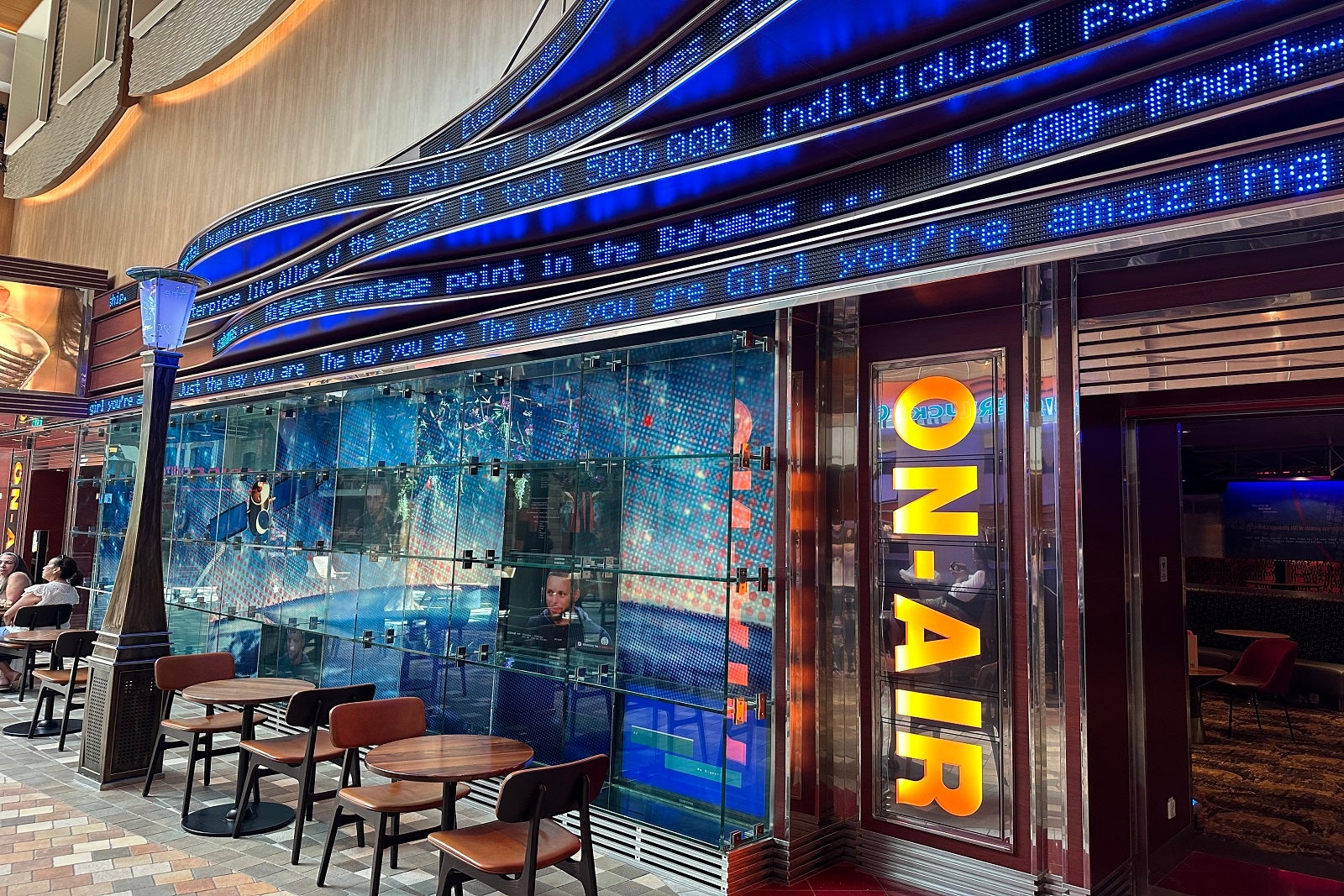 The outside of a cruise ship karaoke bar with tables and chairs near the entrance, a sign that says "On-Air" and song lyrics scrolling by on LED screens