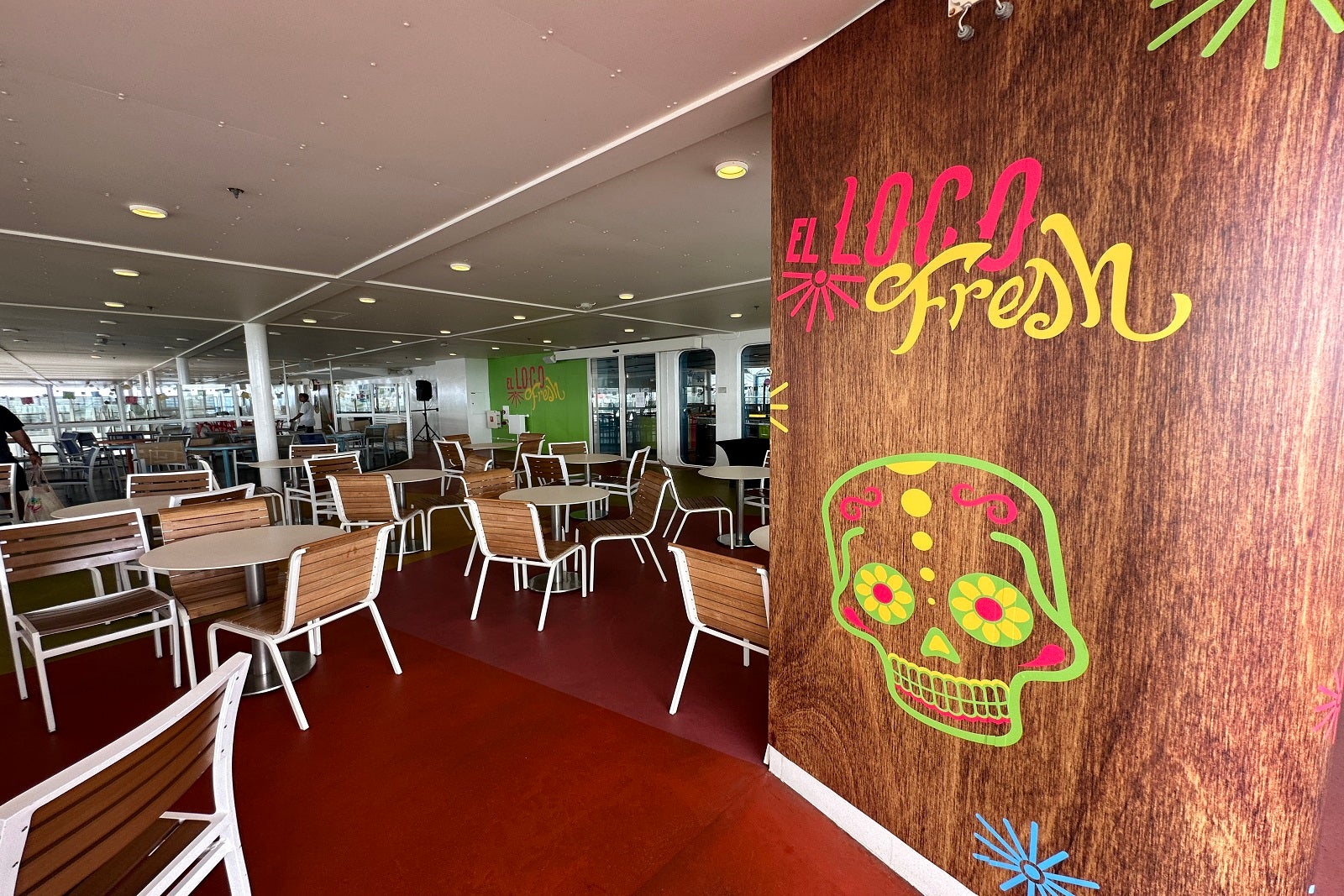 A cruise ship restaurant on an outdoor deck with tables, chairs and a faux wood wall with a sign that reads "El Loco Fresh"