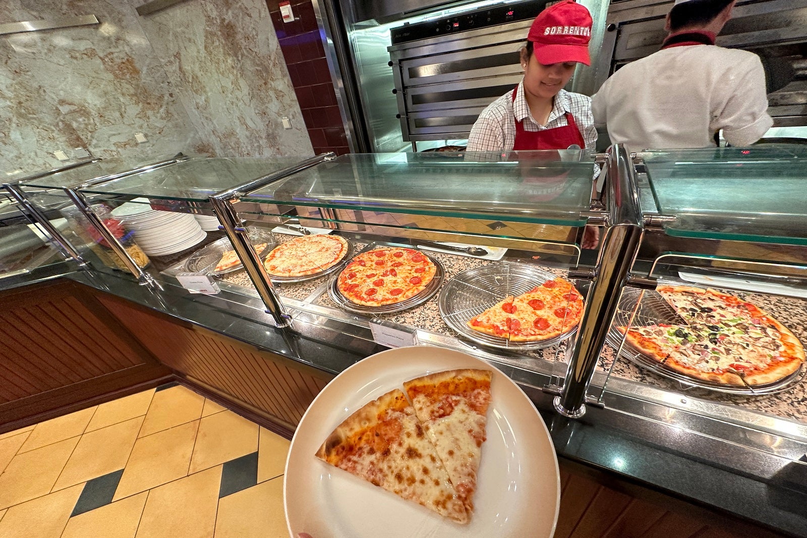 A cruise ship crew member making fresh pizza behind a counter with a white plate and a slice of pizza in the foreground