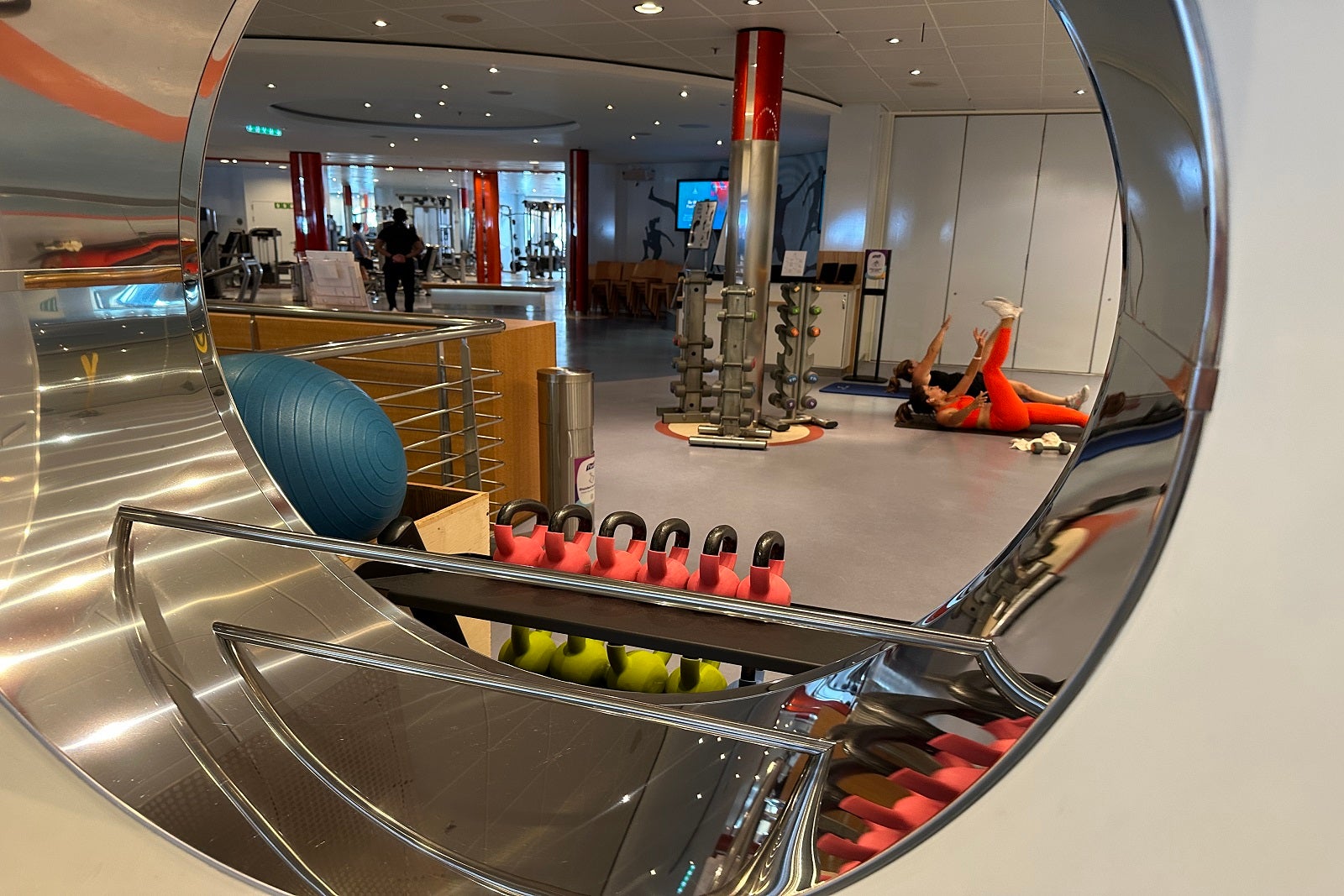 A cruise ship fitness center with kettlebells, exercise balls, weights and two people stretching on the floor