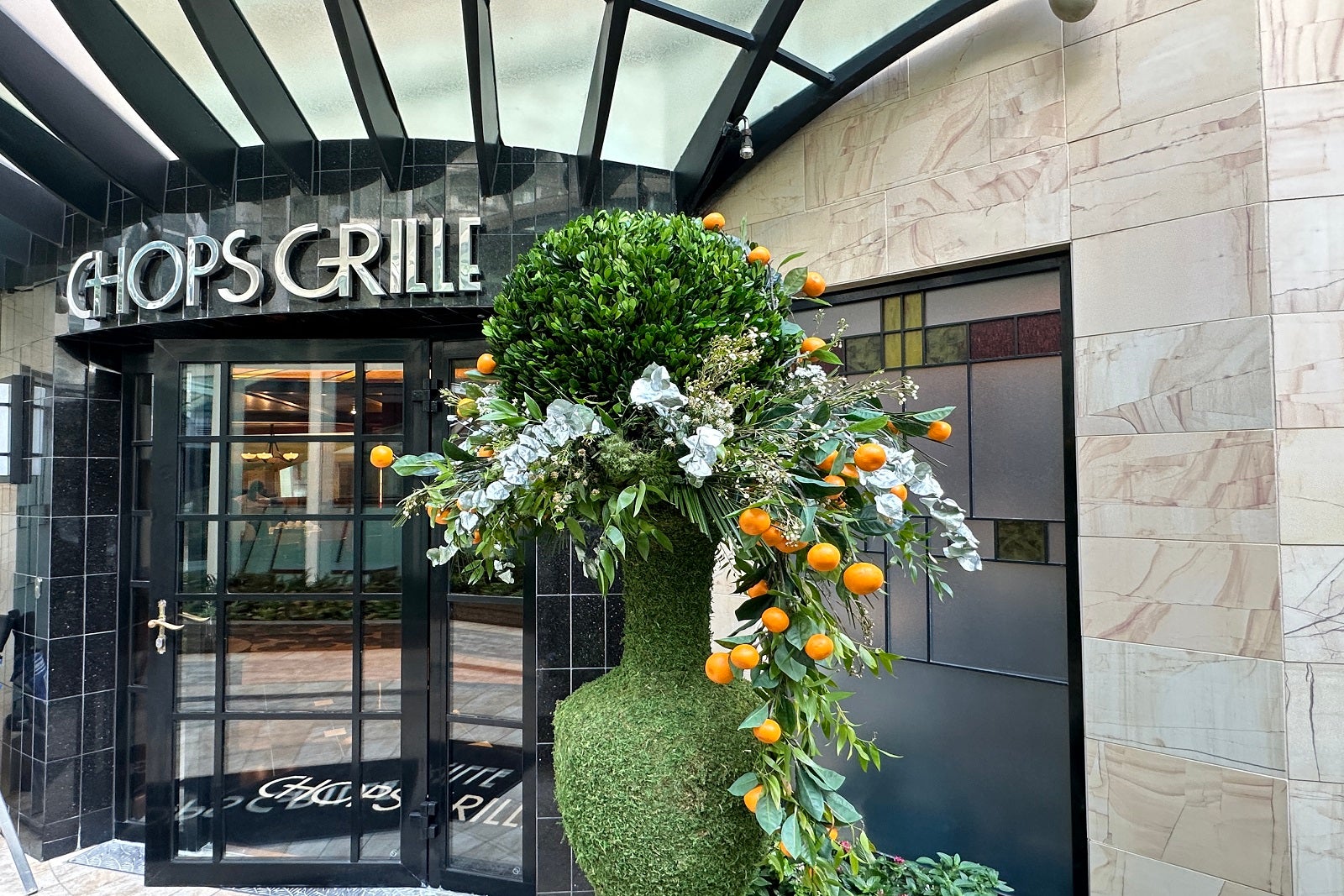 The entrance to a cruise ship steakhouse restaurant with a topiary on one side of the door, a trellis overhead and a sign that reads "Chops Grille" in art deco lettering