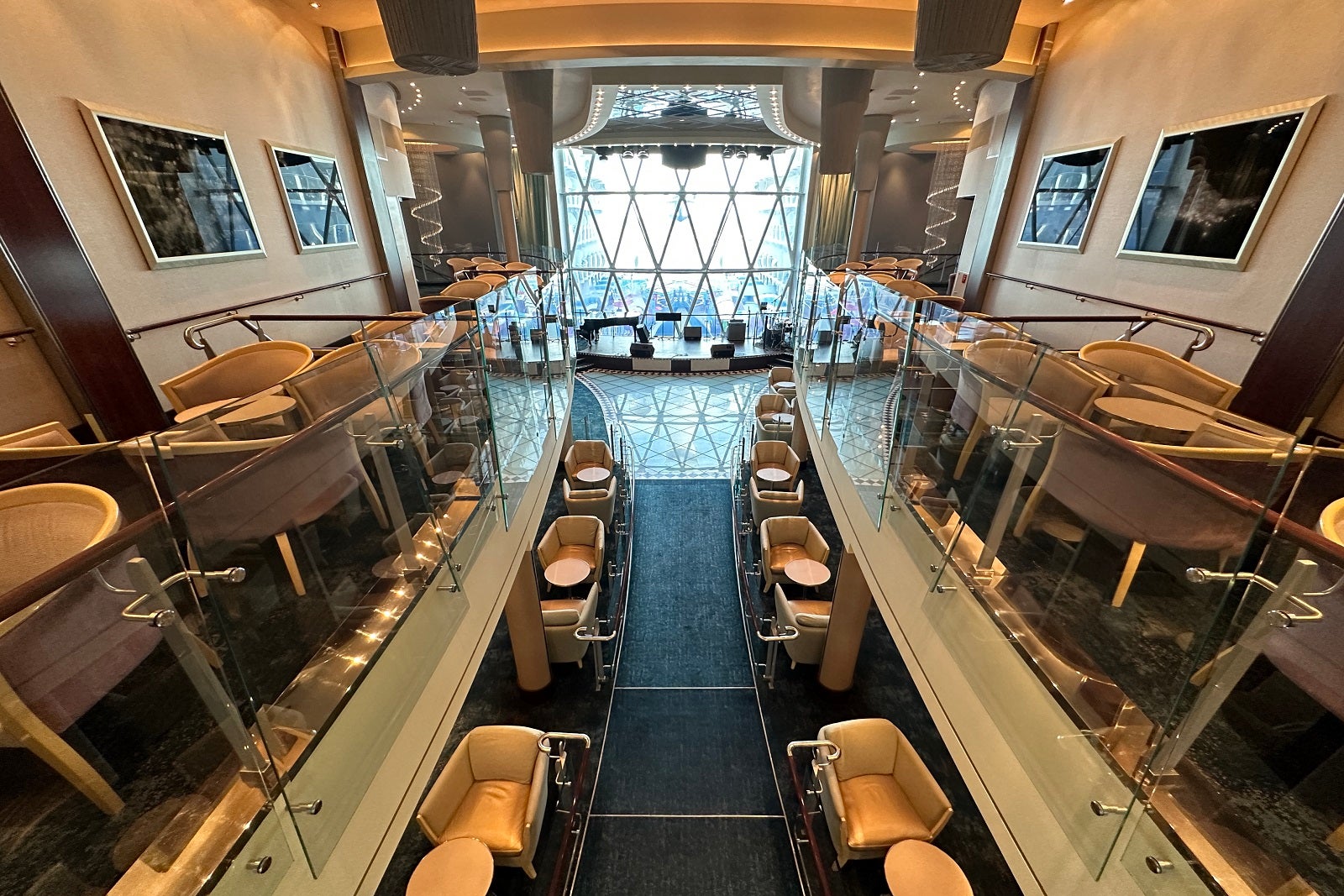 An aerial view of a cruise ship lounge with a stage and dance floor in front of floor-to-ceiling windows with two decks of seating on both sides