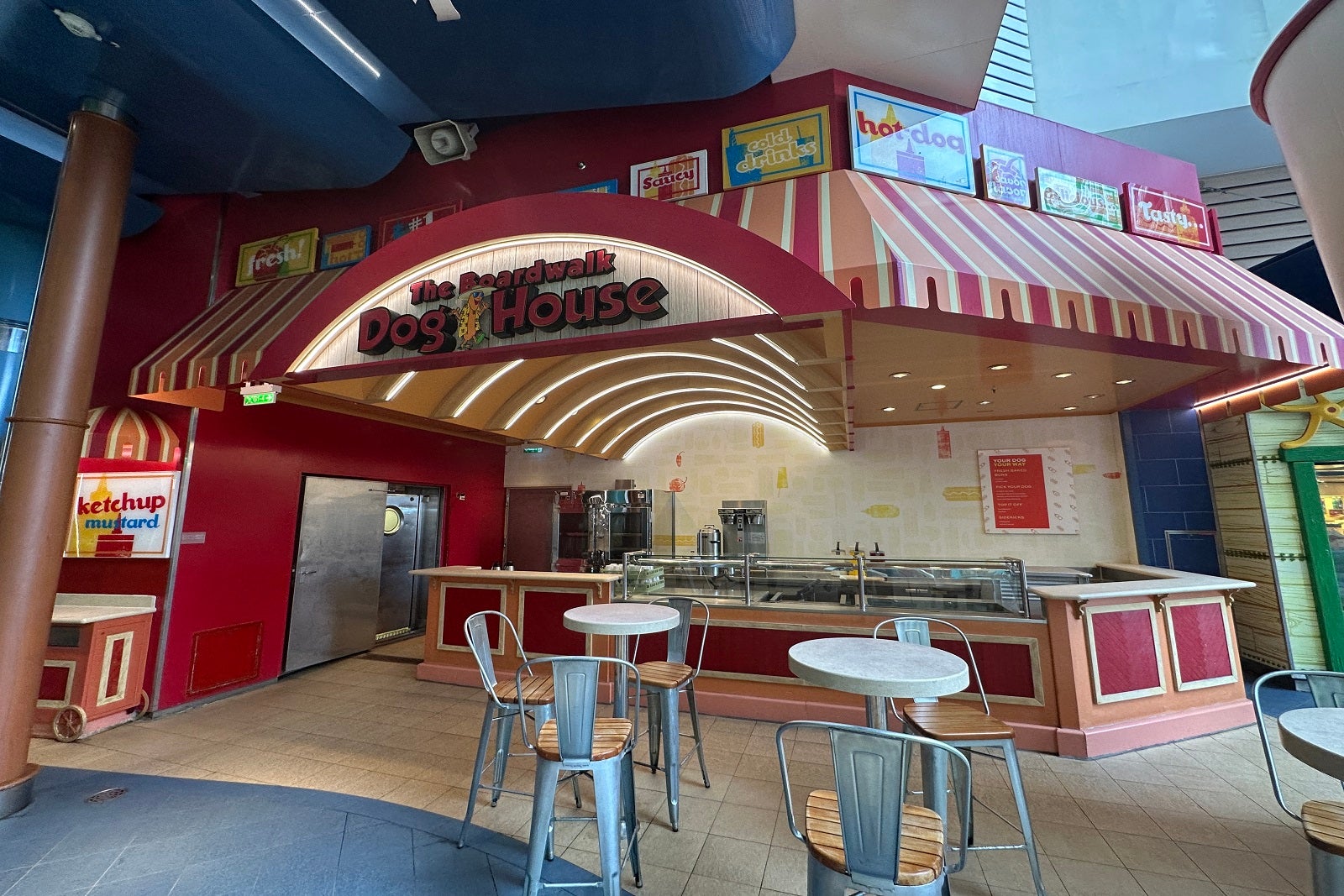 An outdoor boardwalk-style hot dog restaurant on a cruise ship with tables and chairs out front and a sign that reads "Boardwalk Dog House"