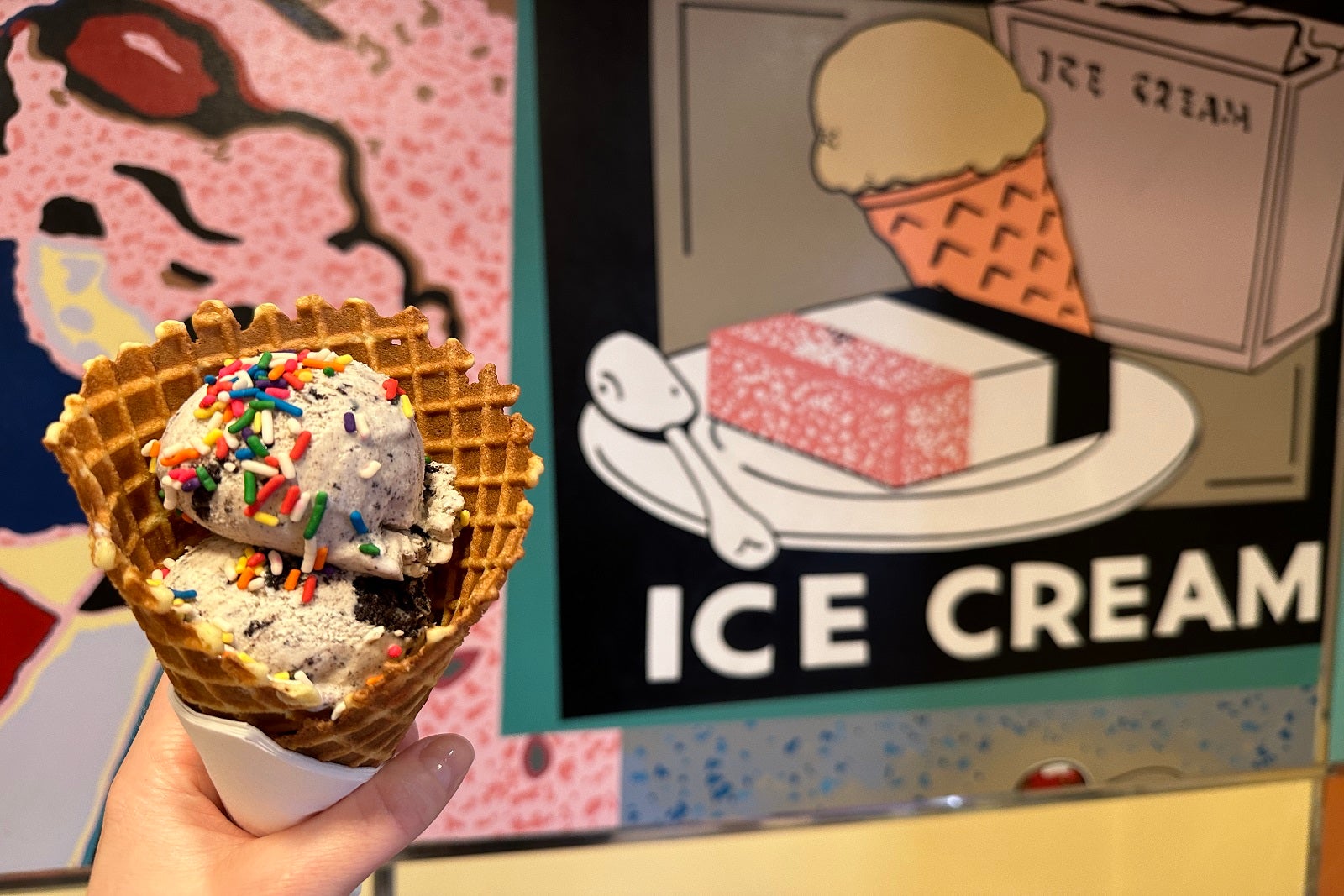 A hand holding a waffle cone with a scoop of ice cream and colored sprinkles in front of a wall that says "ice cream"