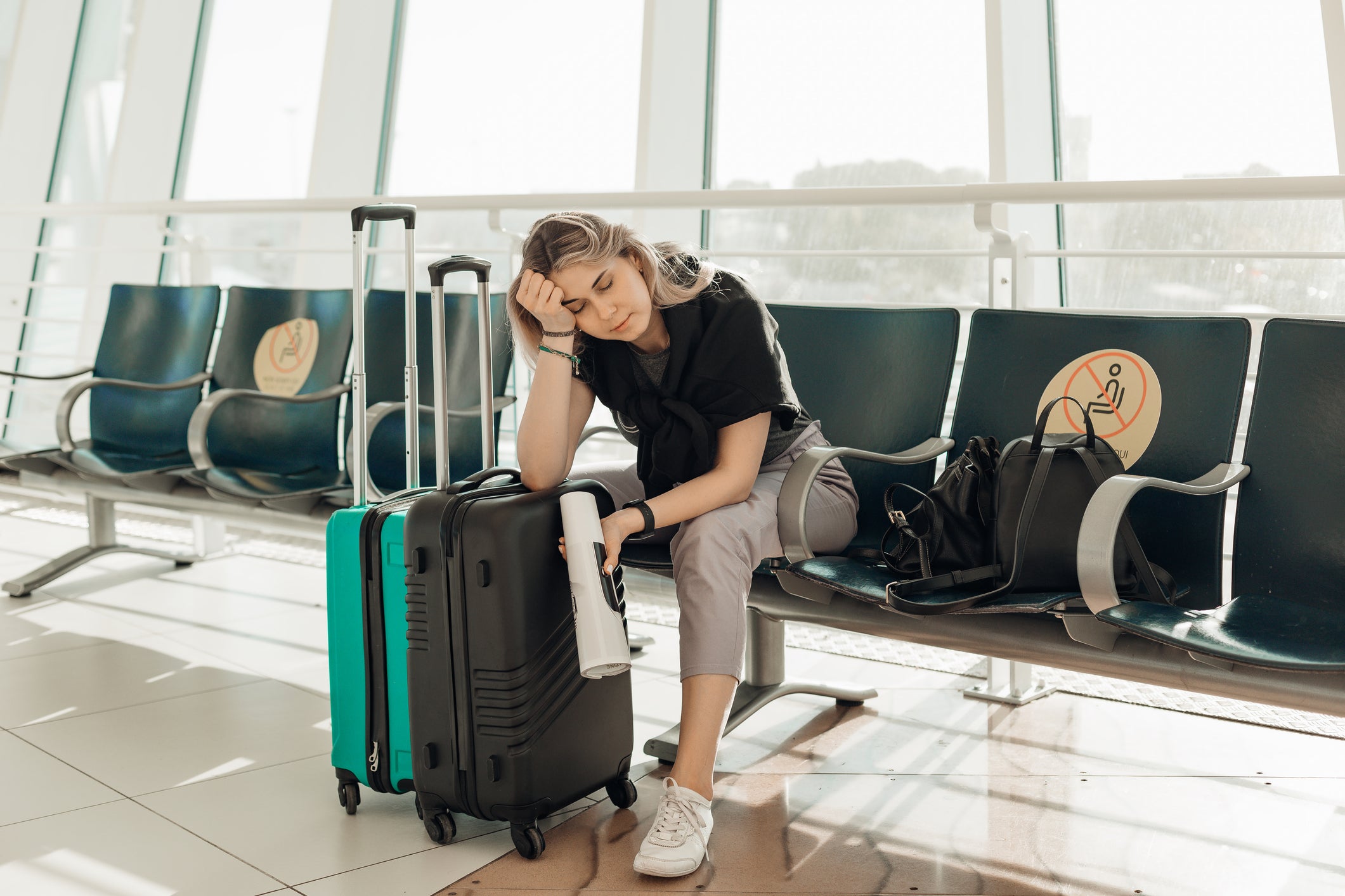 A woman with luggage, leaning elbow on bags, sits in a waiting room at an airport.