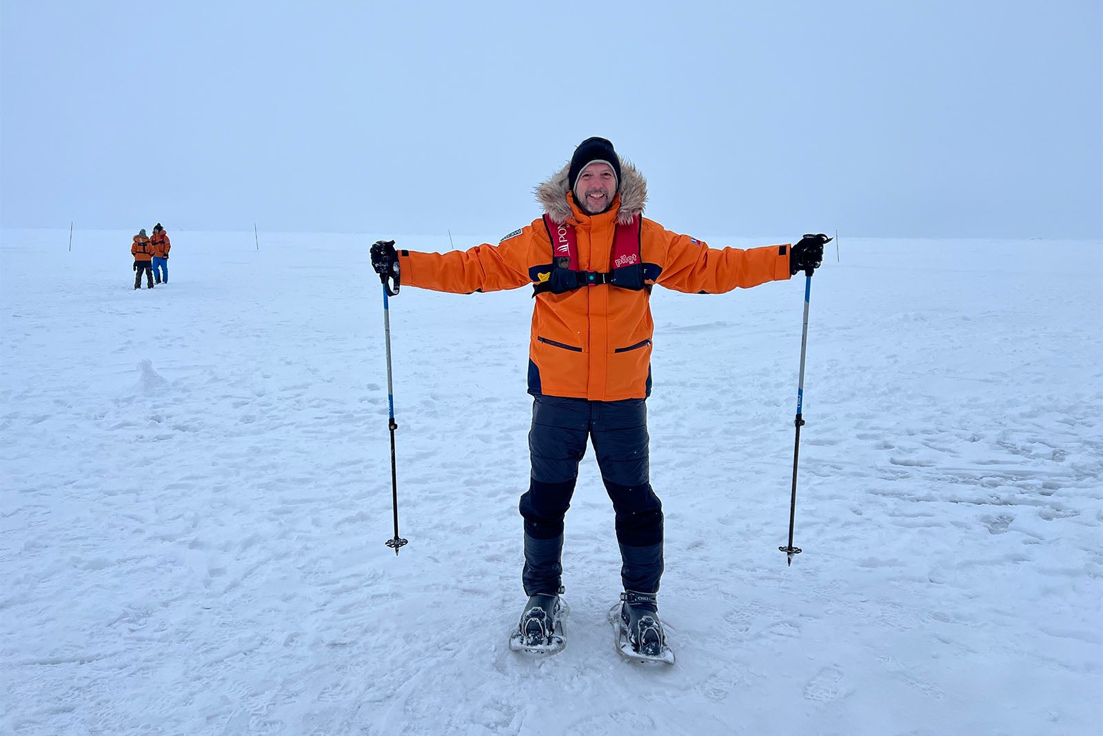 Man in orange jacket and black pants on snow field in Arctic with snowshoes and poles