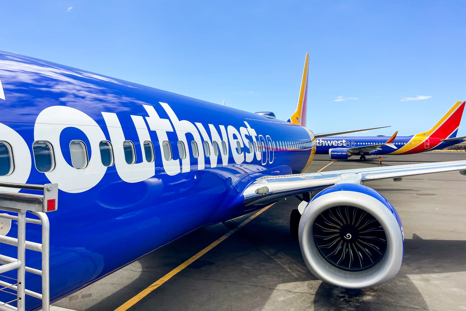 A blue Southwest Airlines Boeing 737 MAX 8 is parked at an airport gate, with another Southwest plane taxiing in the background..