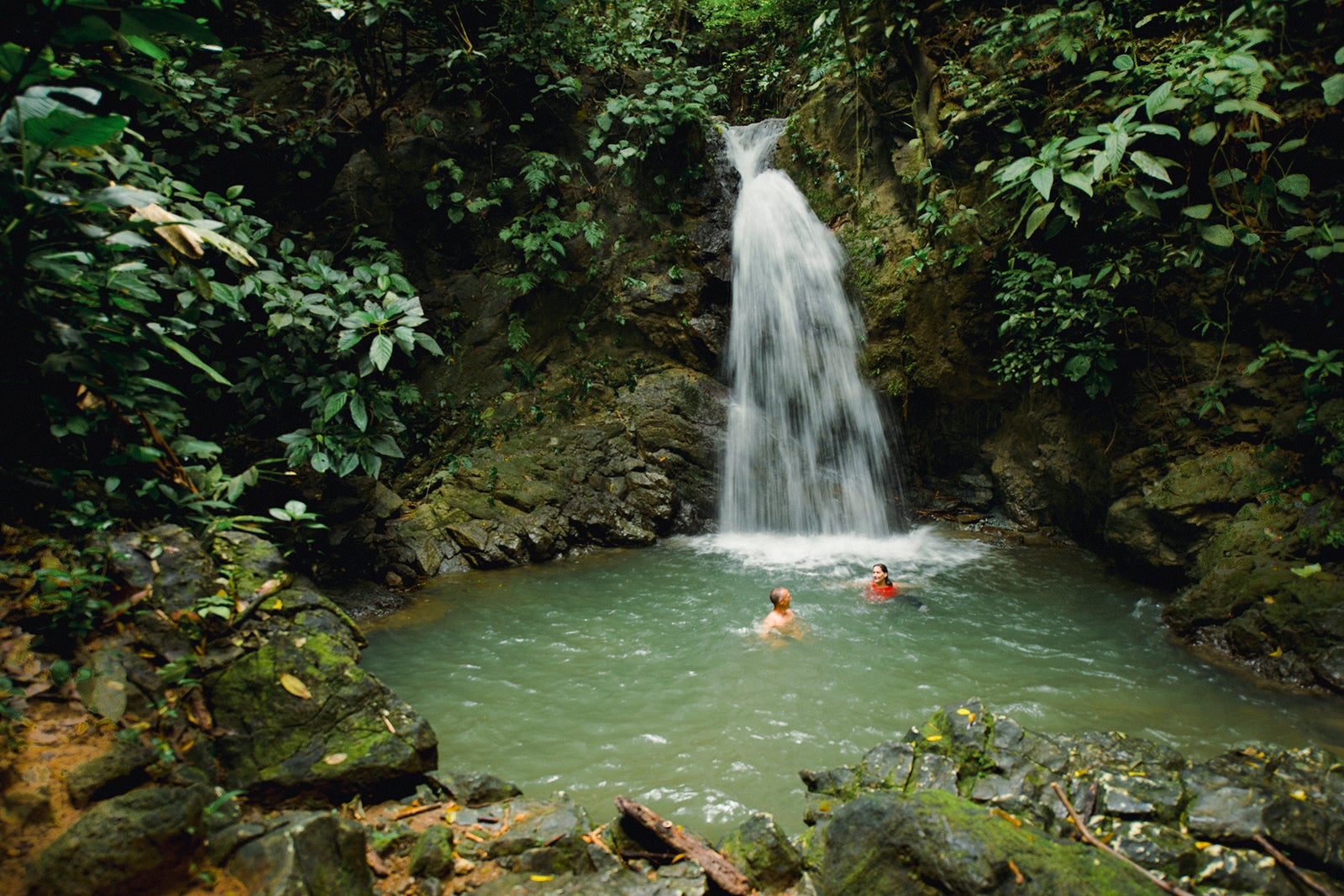 two people swimming in pond beneath waterfall
