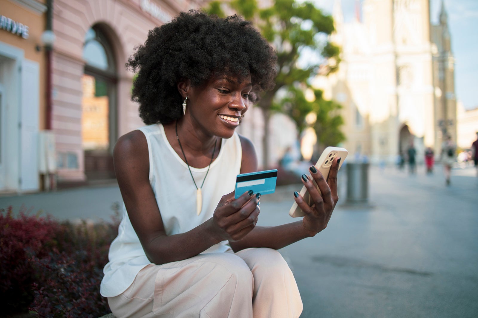 woman sitting on bench in city using smartphone and credit card