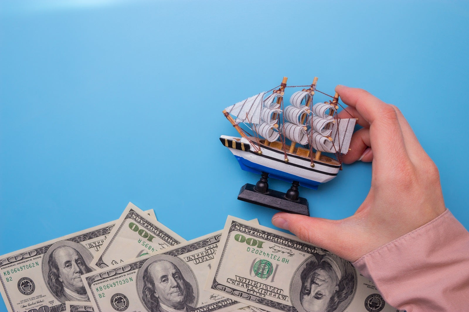 Flat-lay photo of a hand holding a toy sailing ship above a sea of US $100 bills