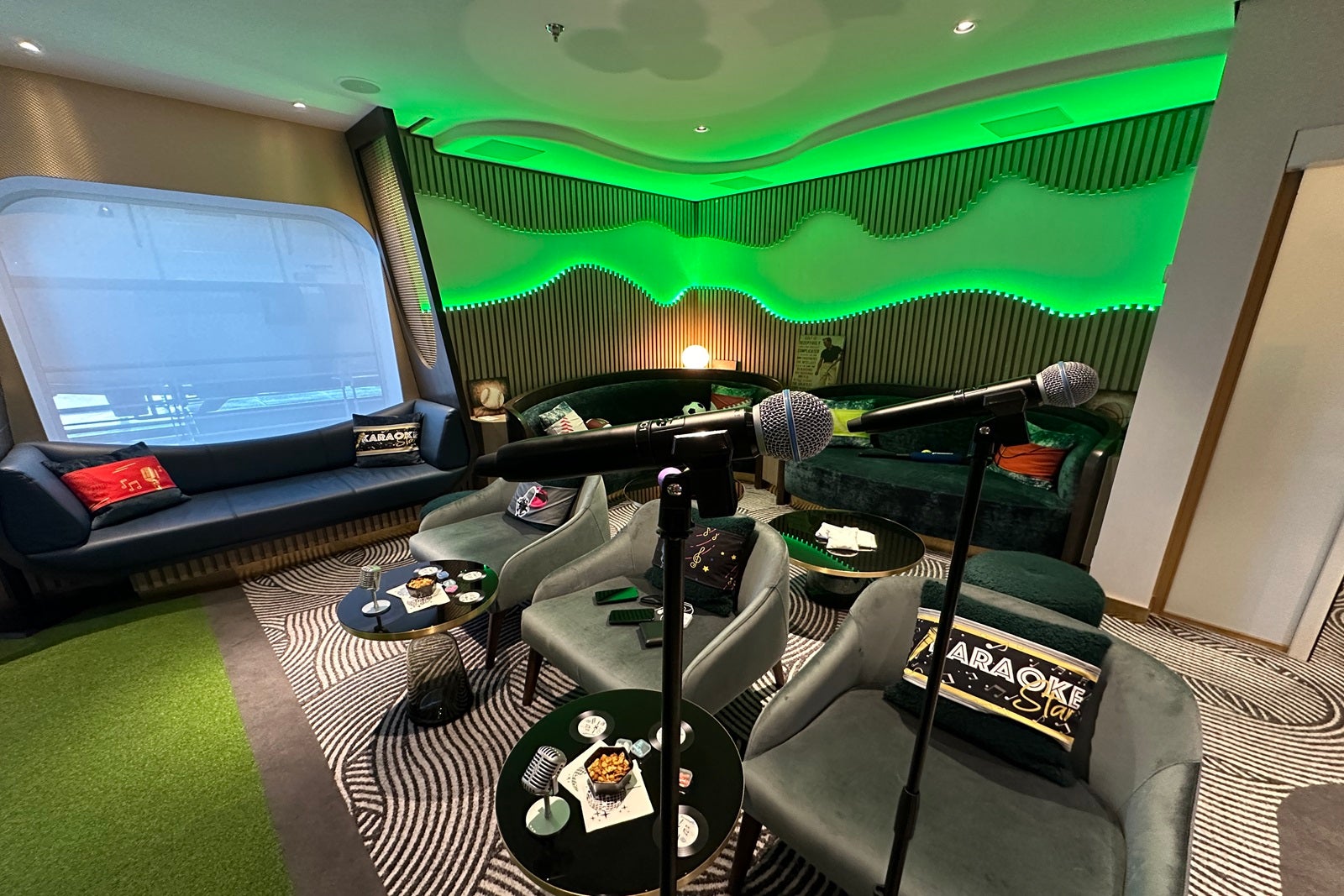 A lounge area with chairs, sofas, throw pillows and funky green lighting. There are also small drinks tables and a couple of microphones for singing karaoke. 