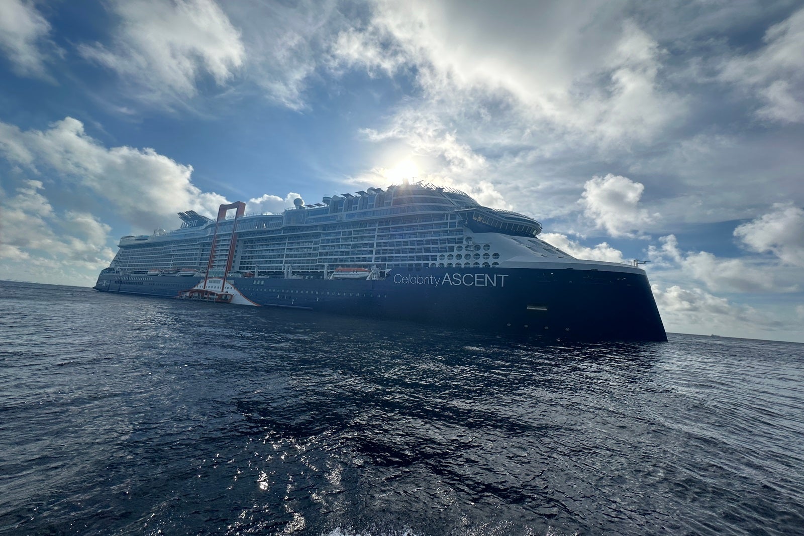 A cruise ship -- Celebrity Ascent -- floats in the water with the sun, blue sky and clouds behind it.
