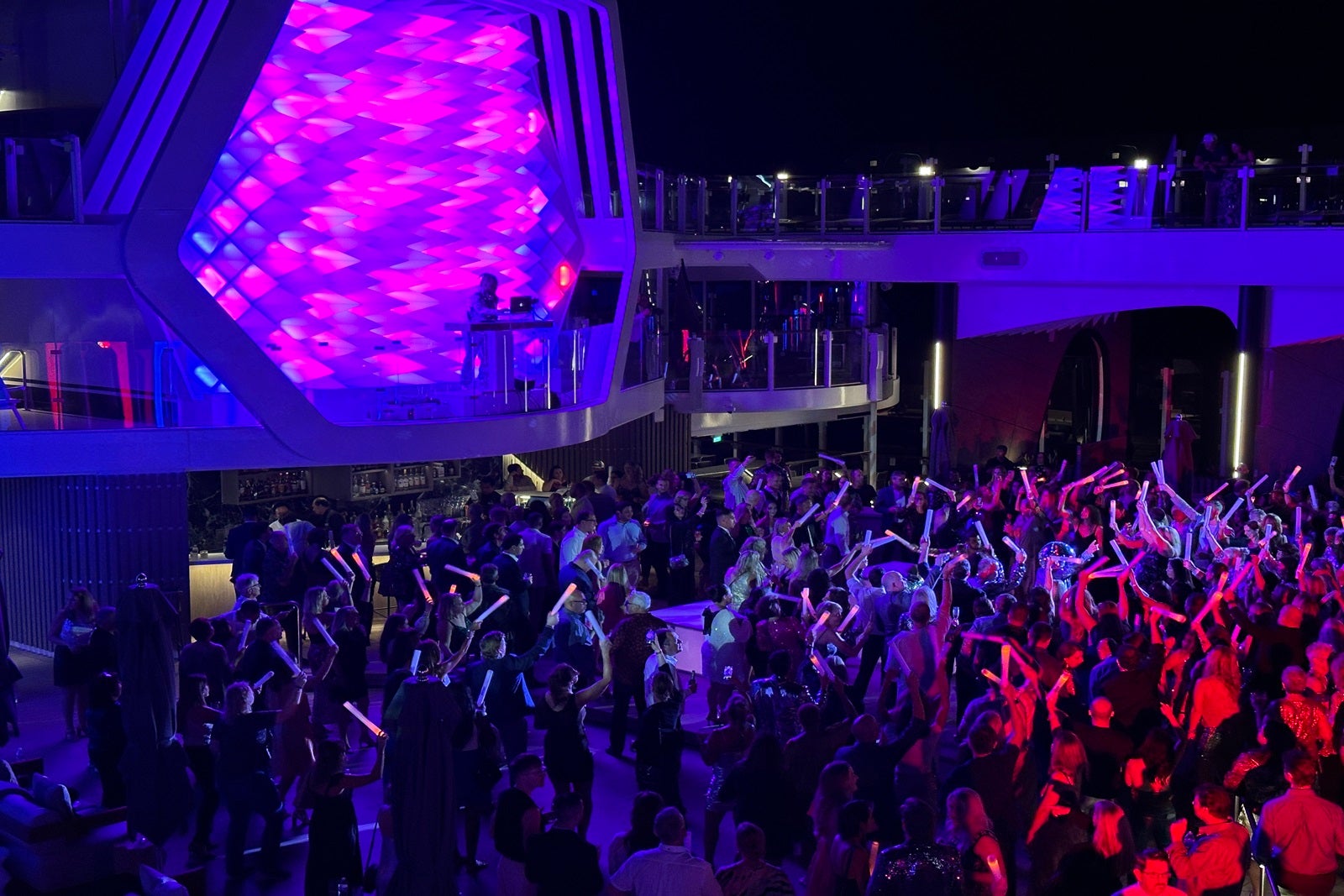 A dancing crowd gathered in front of a DJ booth for a party on the pool deck of a cruise ship at night