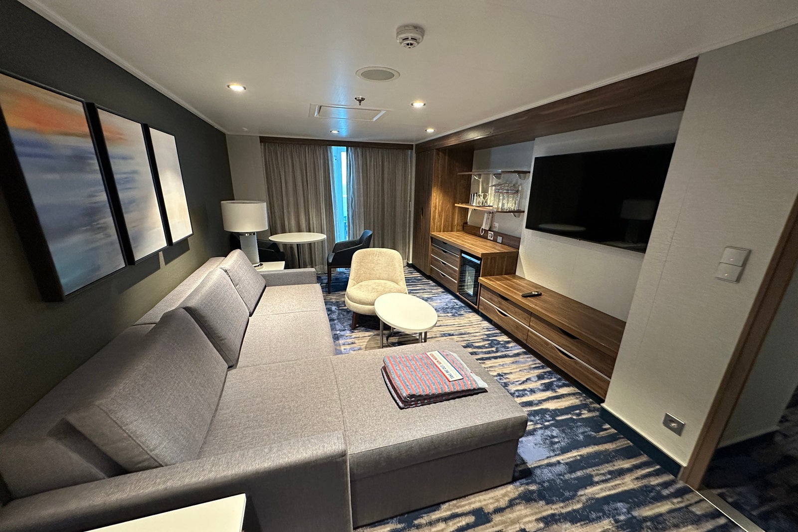 A living room in a suite on a cruise ship with a gray sofa, a table and chairs and a TV