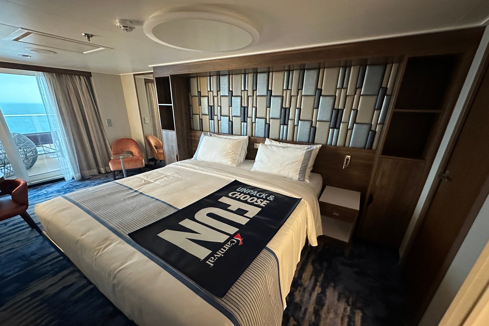 A cruise ship cabin with a bed, a nightstand and a chair with a checkered headboard and a luggage runner that says "Choose fun"