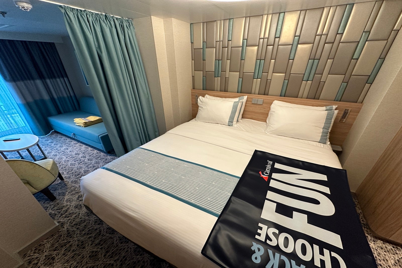 A cruise ship cabin with a bed and a luggage runner that says "Choose fun" with a checkered headboard behind it and a dividing curtain to the side