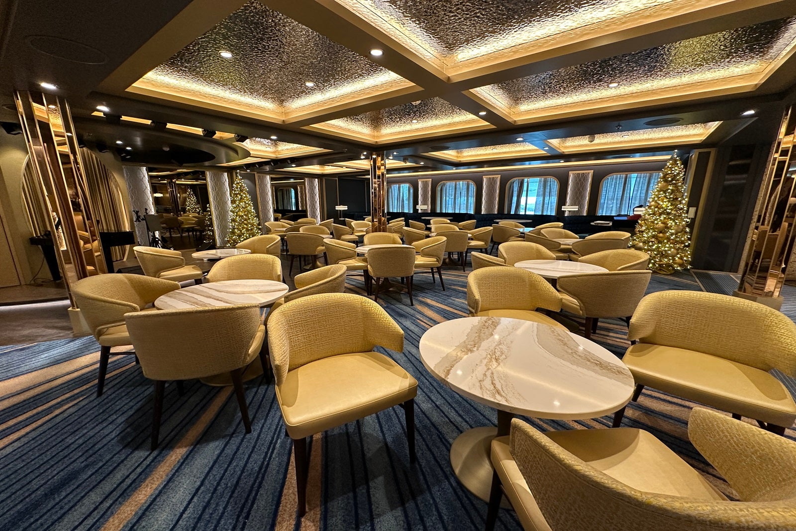 Tables with gold upholstered chairs fill the seating area near a bar on a cruise ship