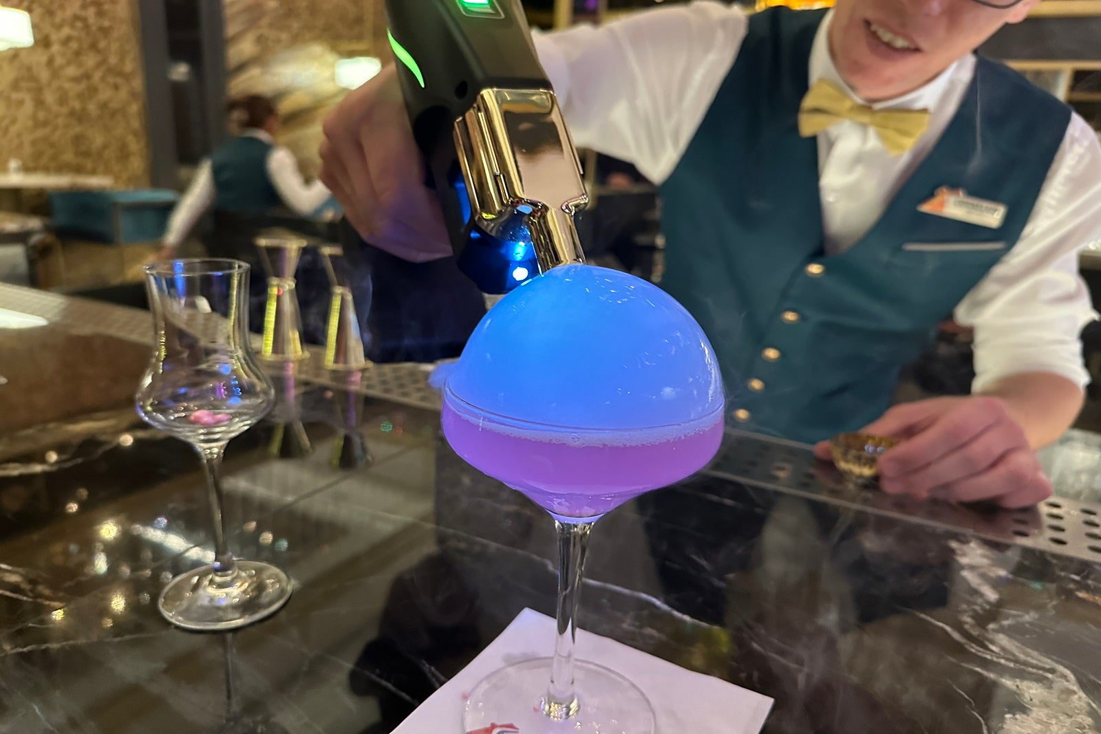 A bartender with a bubble gun putting a smoke bubble on top of a purple cocktail sitting on a bar