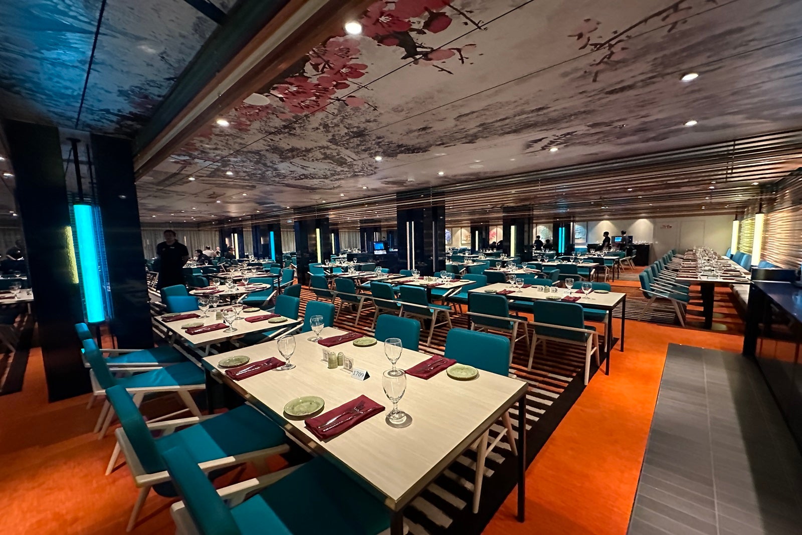 A restaurant with long, rectangular tables surrounded by teal-colored seats on orange carpeting