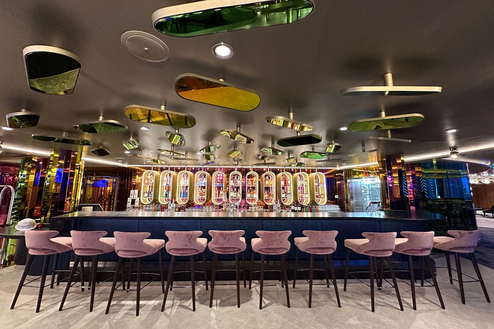 A dark bar with light pink Midcentry Modern chairs, oval-shaped liquor shelves and oveal multi-colored glass elements overhead