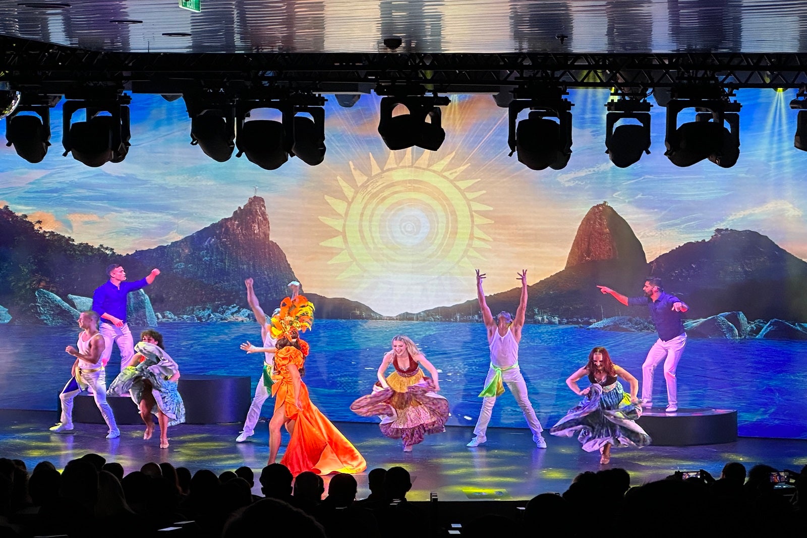 Performers dance to Brazilian music as part of a cruise production show with mountains, water and a sun on an LED screen in the background