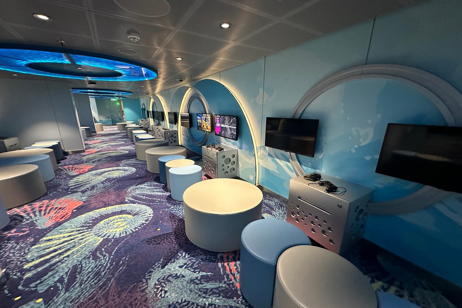 A large carpeted room with cyllindrical stools set around multiple TVs with video game stations