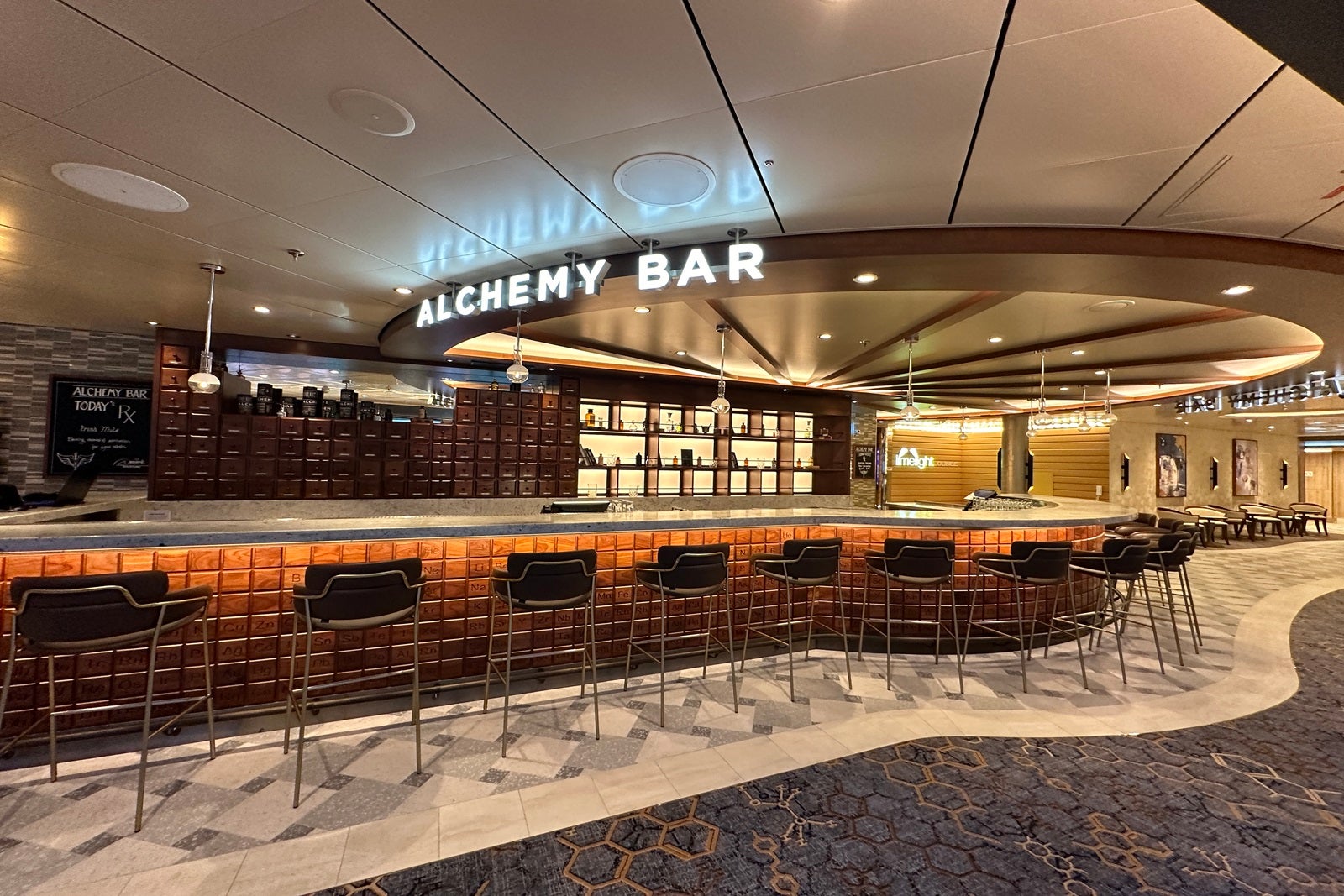 A long, wooden bar with high stools, a backlit set of cubbies holding apothecary paraphernalia and a sign reading "Alchemy Bar"