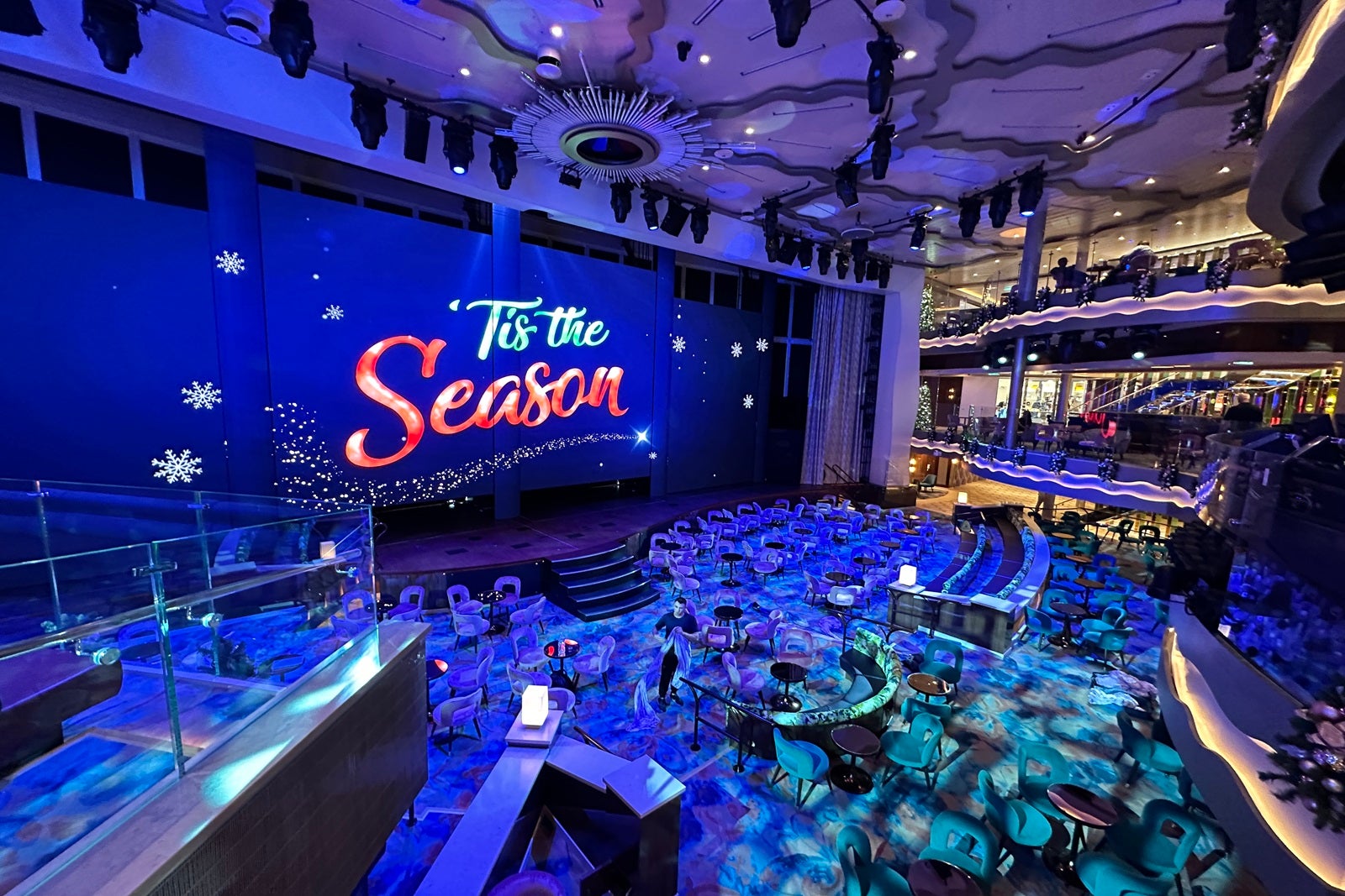 A cruise ship atrium's LED screen displays a "'Tis the Season" holiday message