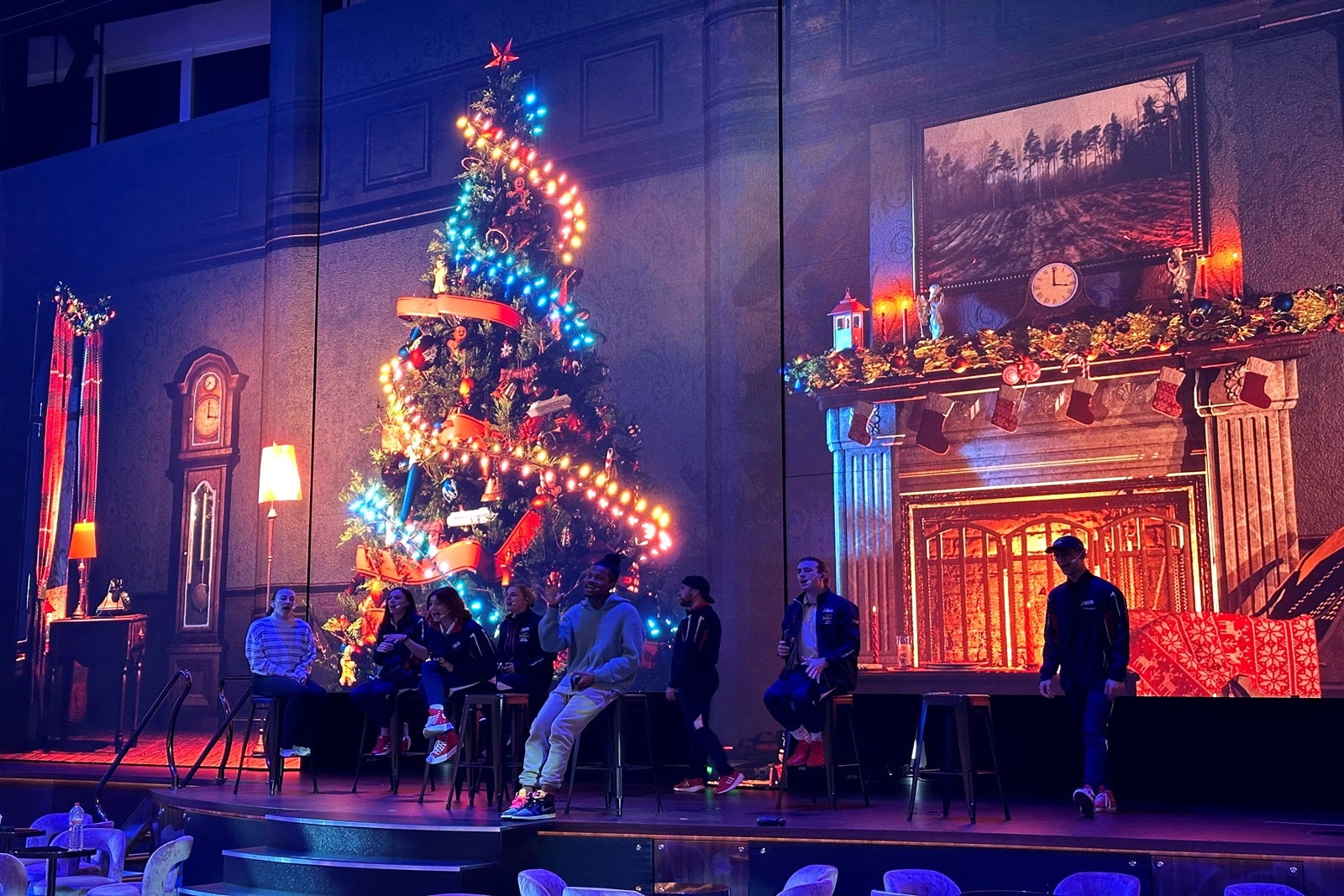 Singers sit on a stage rehearsing with a fireplace and Christmas tree on an LED screen in the background