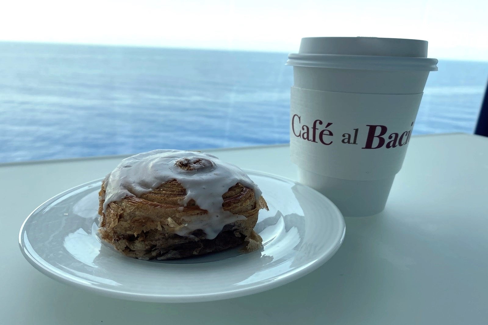A pastry on a dish and a cup of to-go coffee on a table in front of the water on a cruise ship balcony