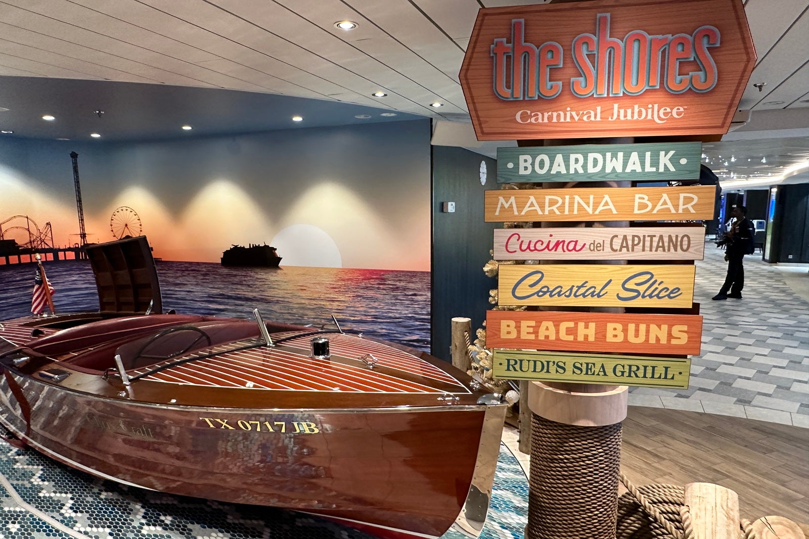 A wooden boat set up as a photo spot with jute-wrapped poles in front of it, a directional sign that shows passengers where different areas of the ship are and a sunset water backdrop behind it