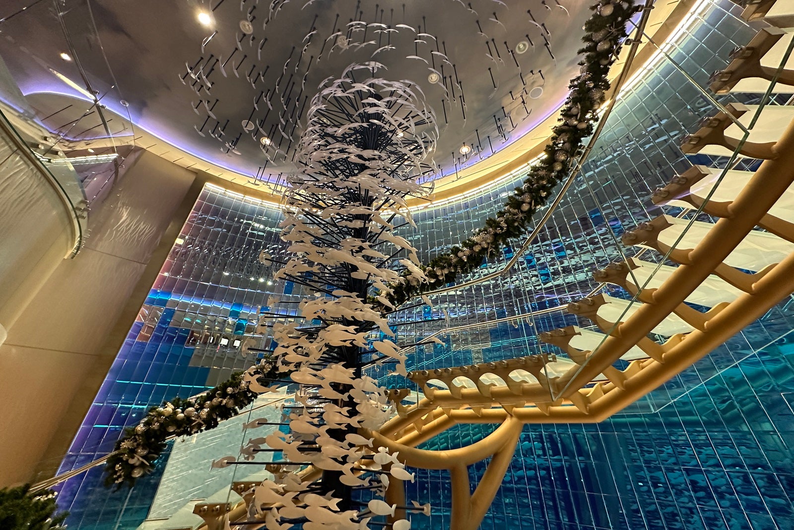 A wall with shiny blue mirror tiles with a golden staircase in front of it and a sculpture of white swimming fish swirling down from the ceiling