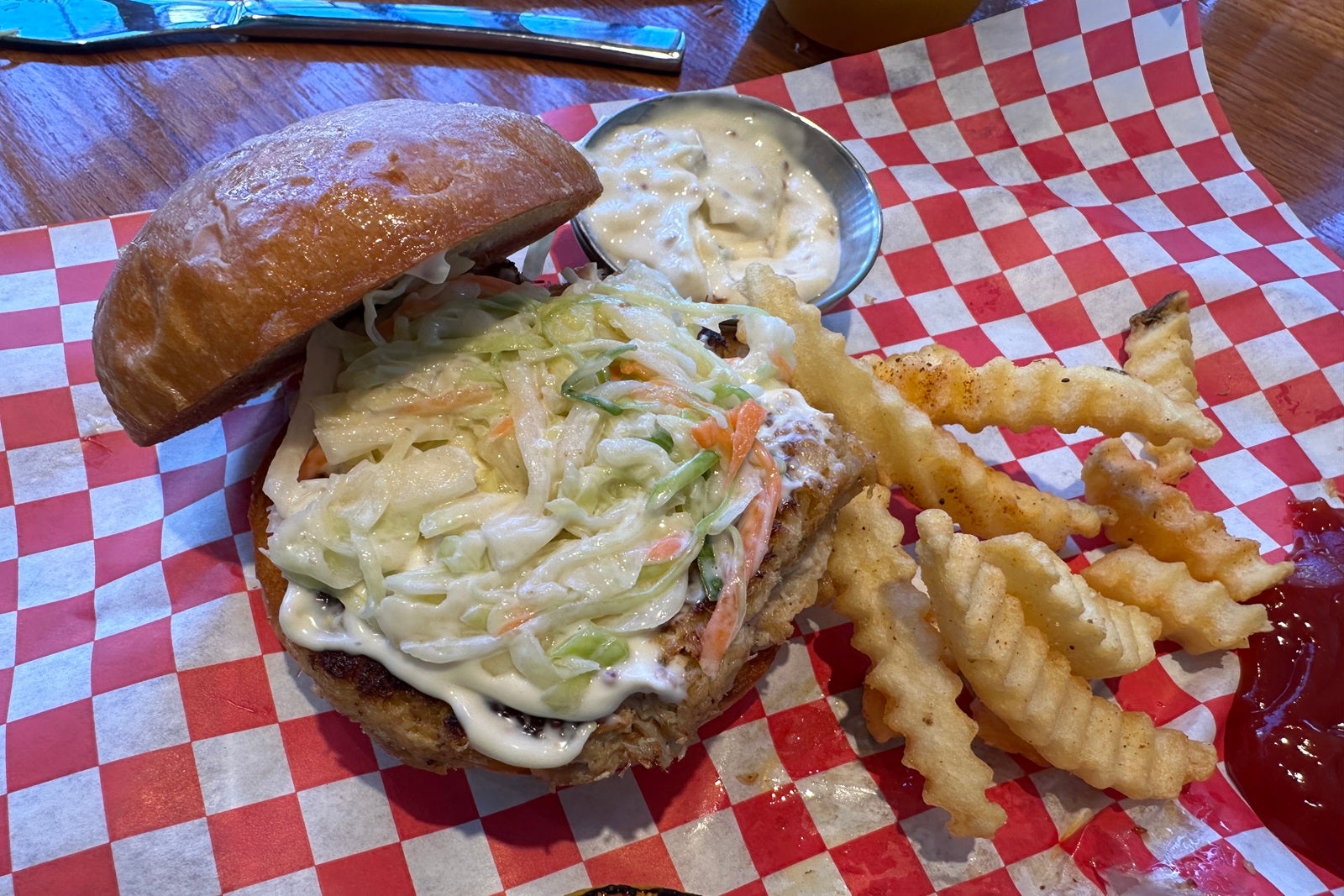 A crab cake topped with coleslaw on a bun with fries next to it on red and white checkered paper