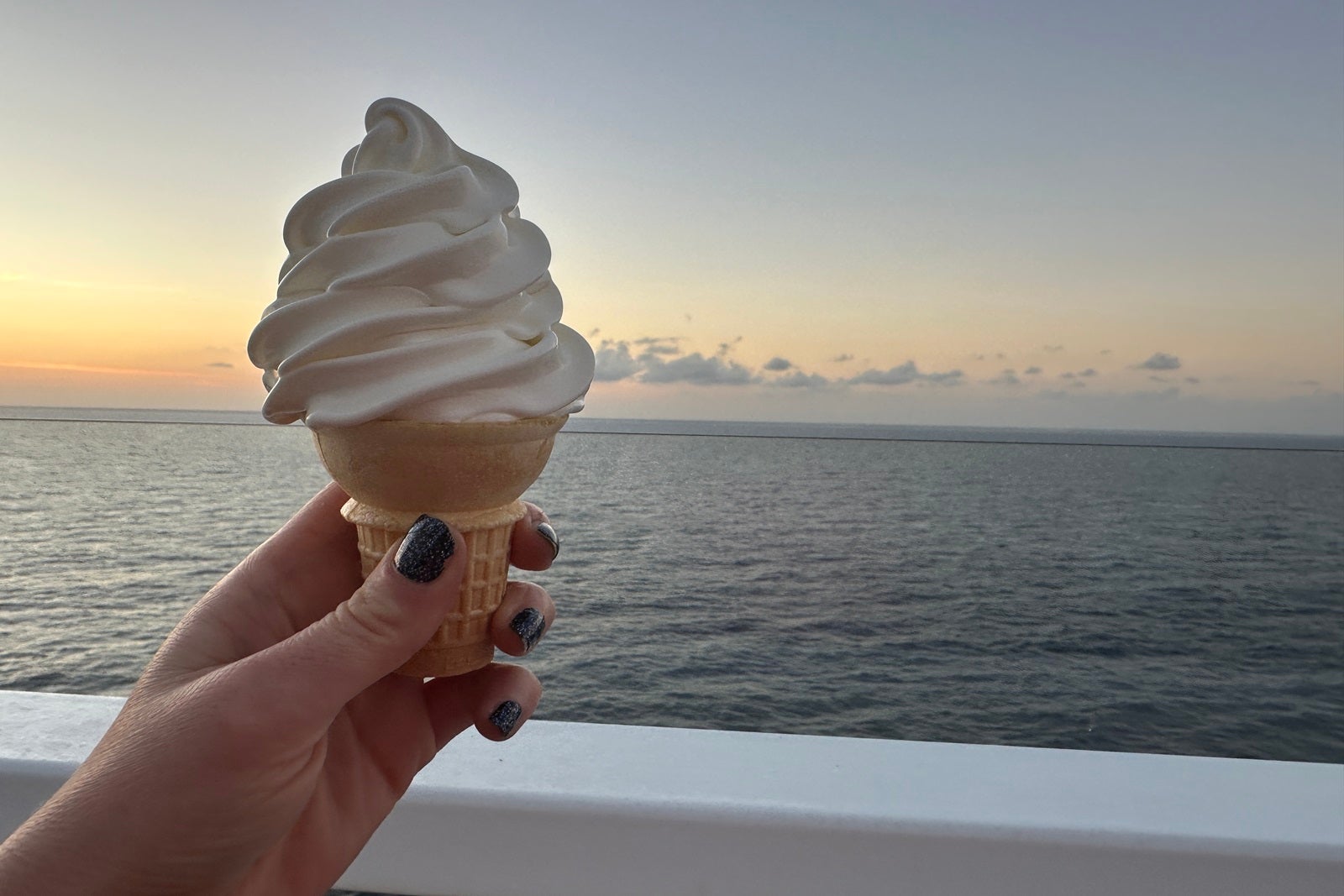 A hand holding a cone of soft-serve ice cream in front of an ocean sunset