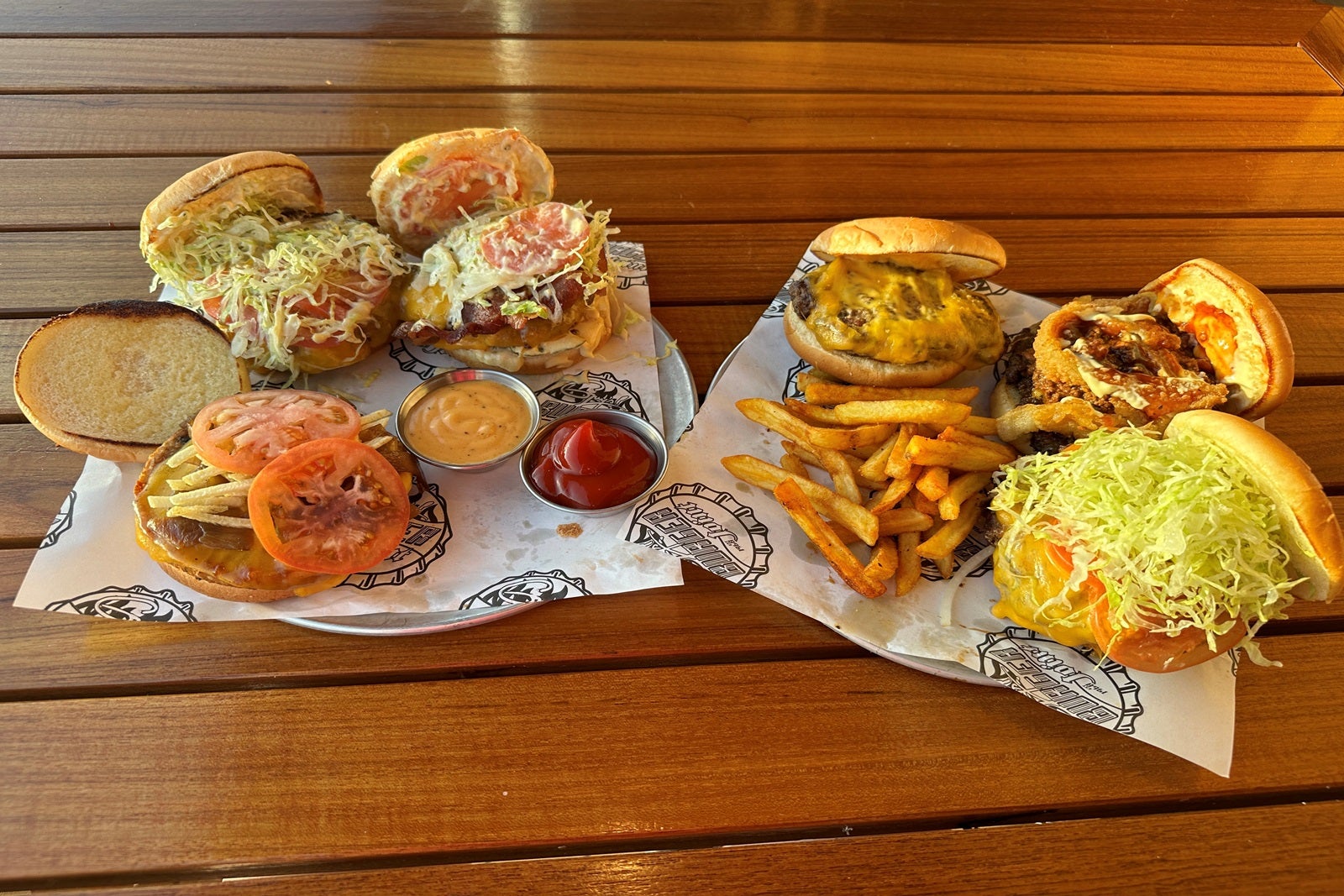 Two sets of three loaded cheeseburgers with fries on two plates on a wooden table