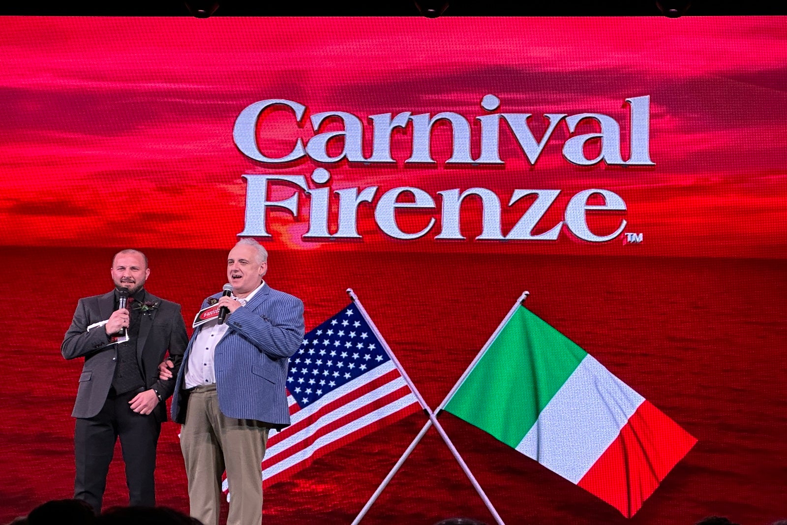Two men on a stage in front of a giant screen that reads "Carnival Firenze" with American and Italian flags under the text