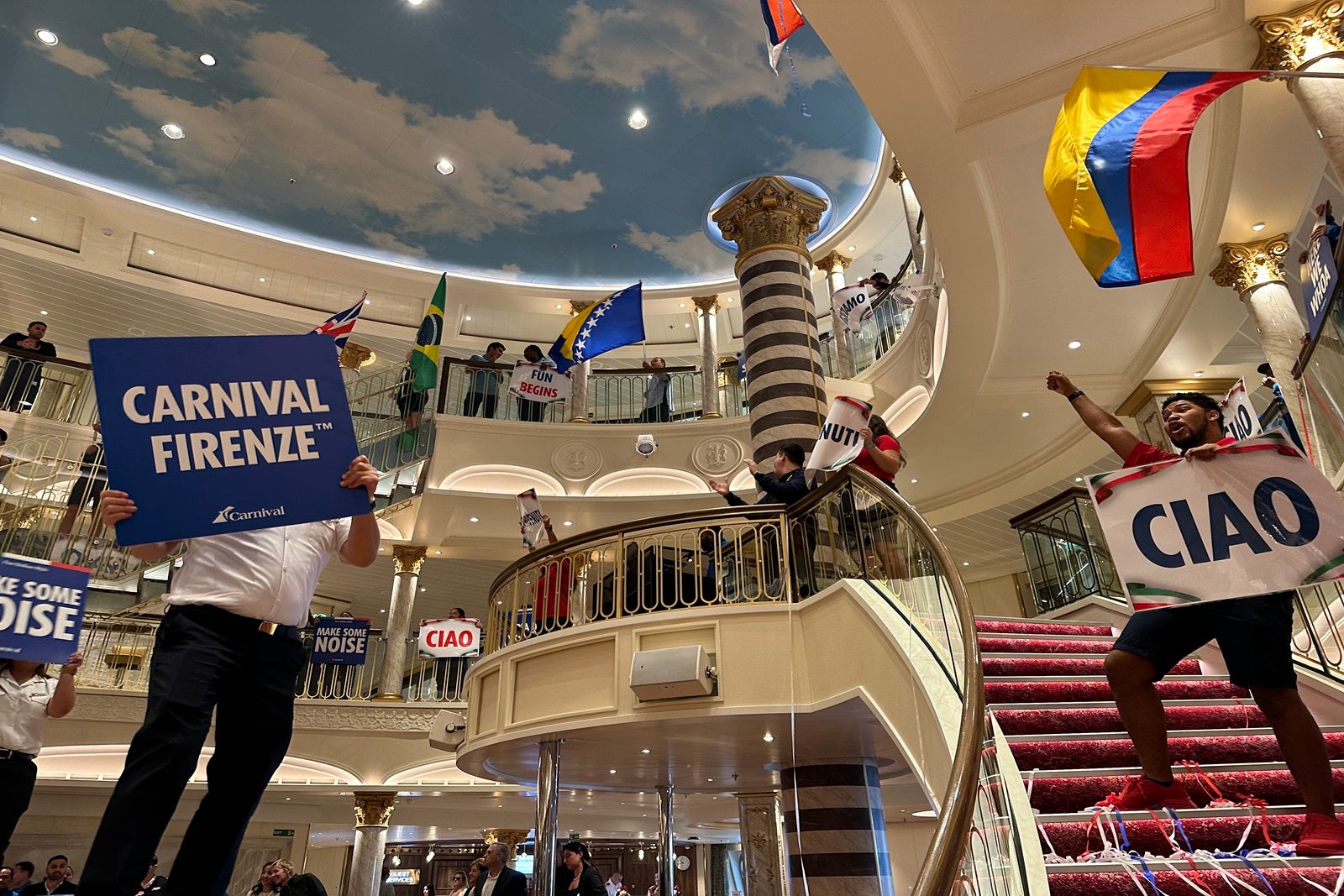 Crew members on a cruise ship wave flags and signs in the atrium to welcome passengrs on board