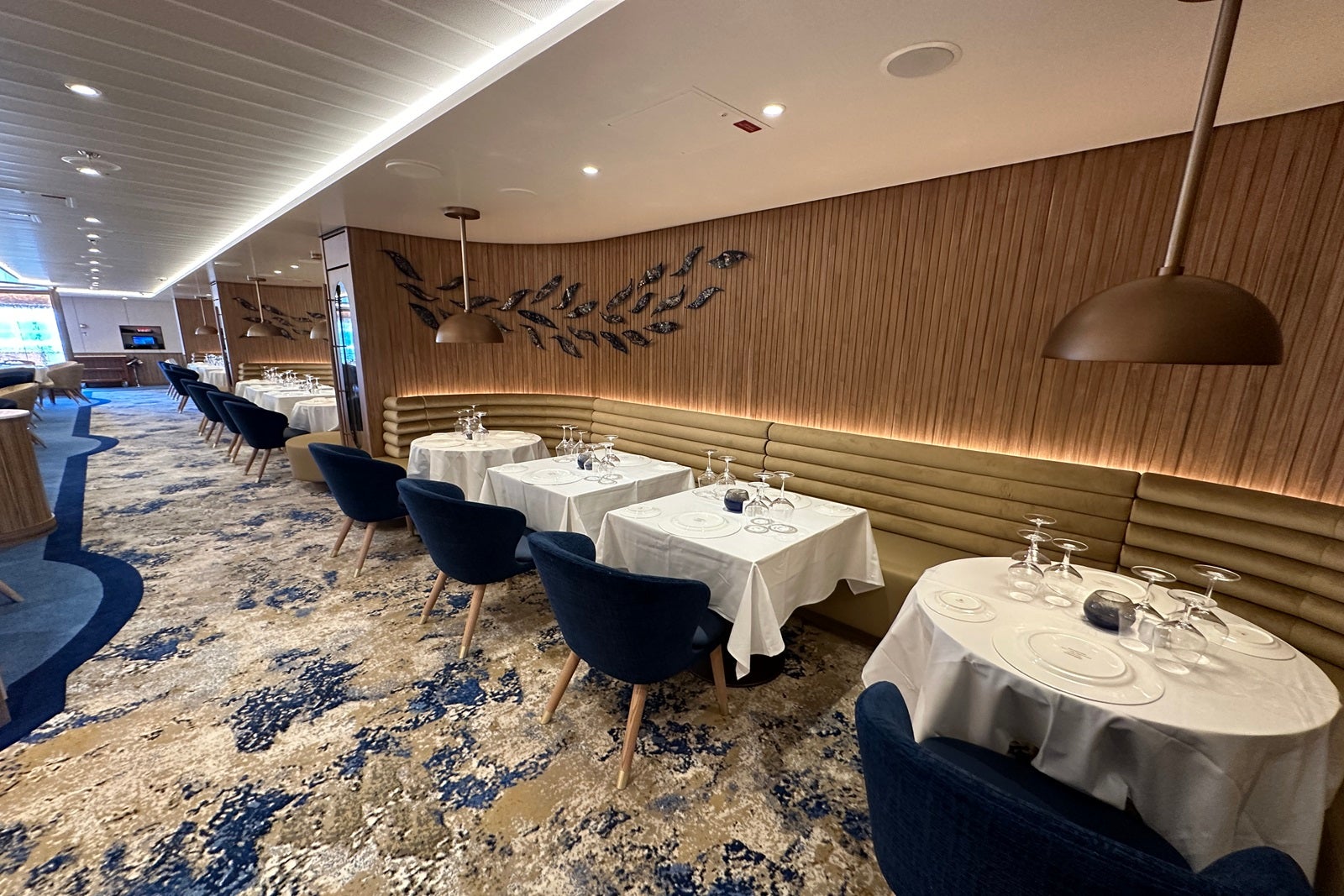 A restaurant with tan and blue carpeting, tables with white tablecloths, blue chairs and a curved, wooden wall with fish designs