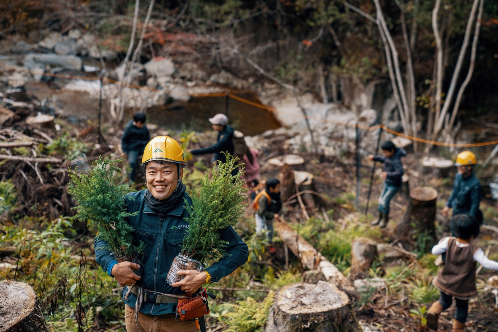 A man in a yellow hard hat carrying two trees to plant in the forest