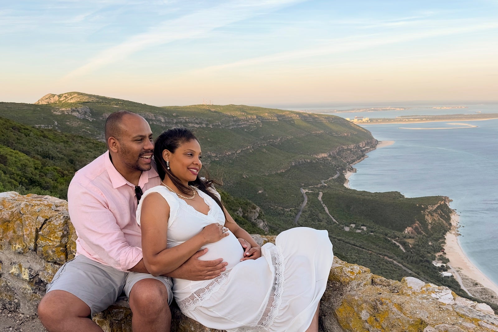 An expecting couple on a babymoon sitting on a rock overlooking the water